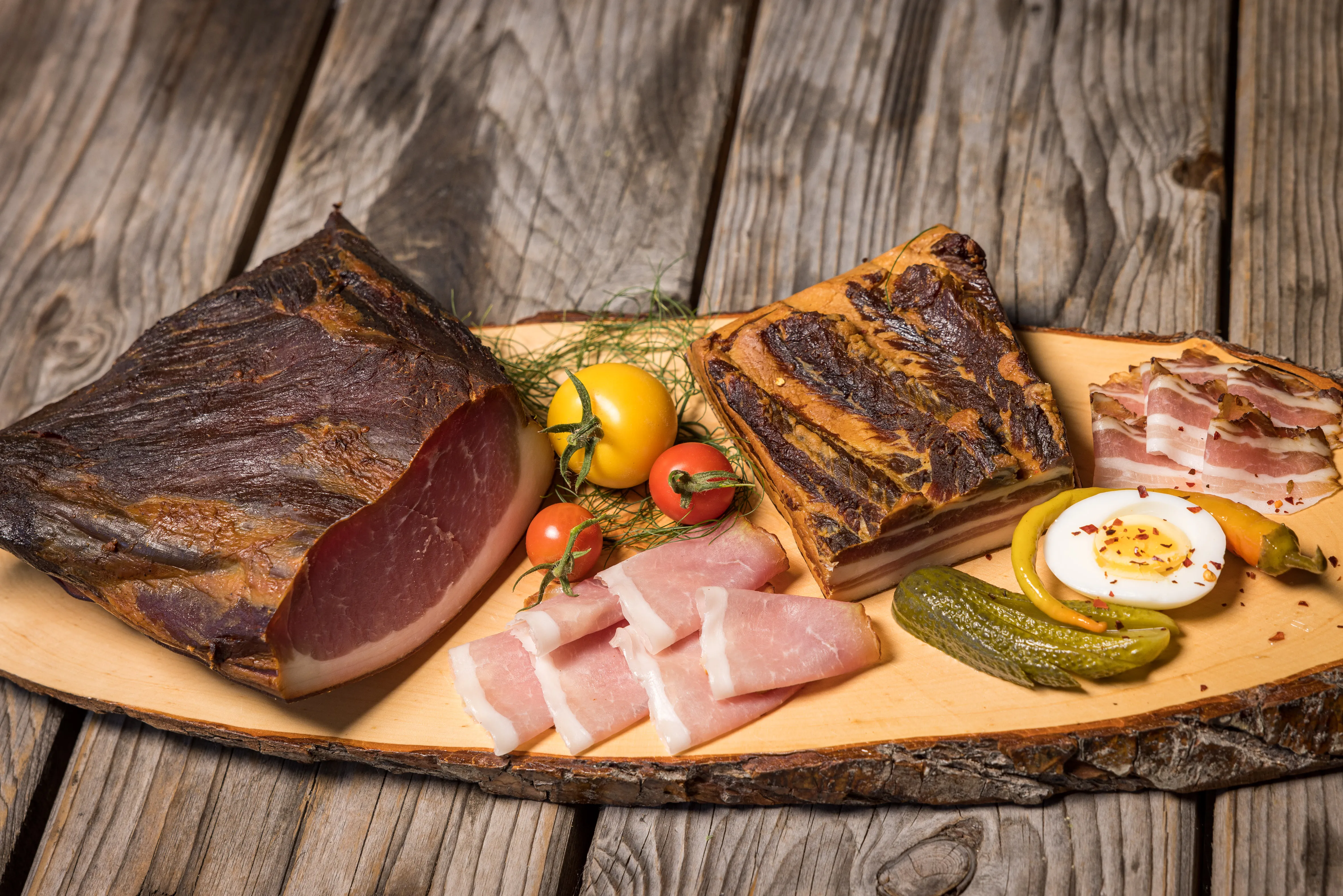 Auf einem Holztisch liegt ein Jausenbrett mit Speck im Ganzen und aufgeschnitten; garniert mit Gurkerl, Ei und Tomate