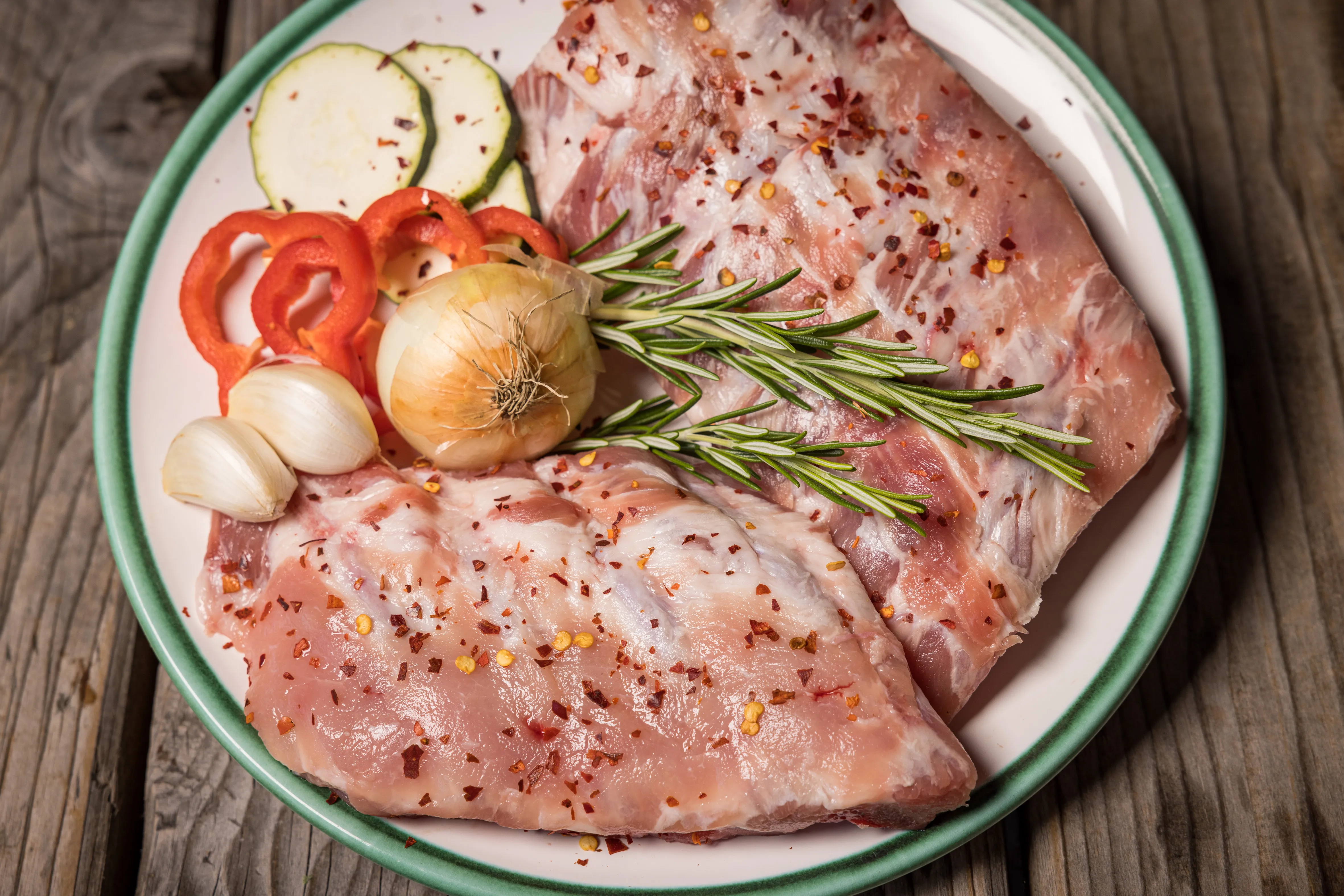 Auf einem Holztisch steht ein Teller mit Frischfleisch, garniert mit Knoblauch, Zwiebel, Paprika, Gurke und Rosmarinzweigen