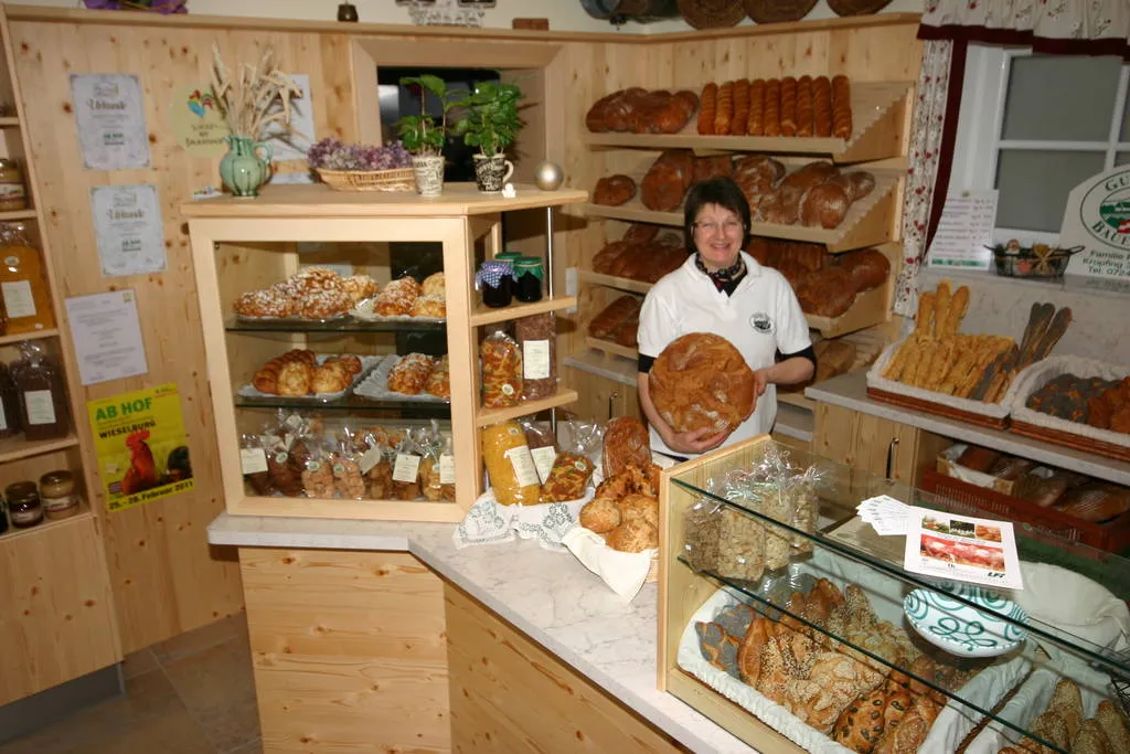 Frau Riedlbauer steht in einem Hofladen hinter einer Theke mit Glasvitrinen. Sie hält eine Laib Brot in der Hand. Dahinter Regale an der Wand mit verschiedenen Produkten. 