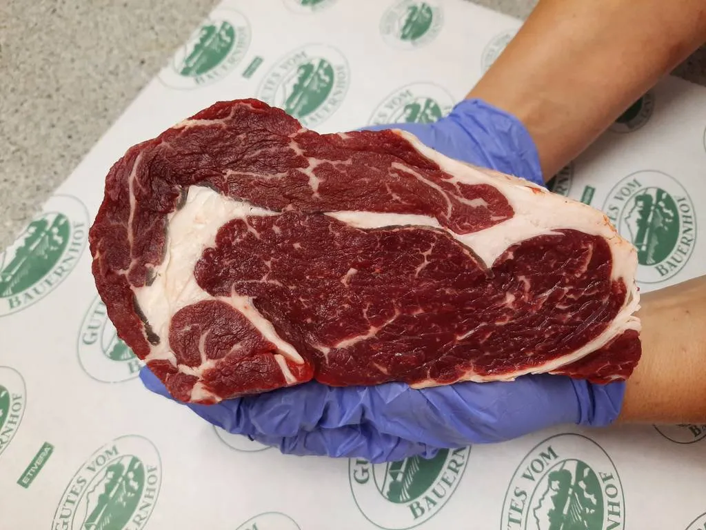 Ein Händepaar mit blauen Handschuhen hält ein großes Stück Fleisch in den Händen; darunter Papier mit der Aufschrift Gutes vom Bauernhof