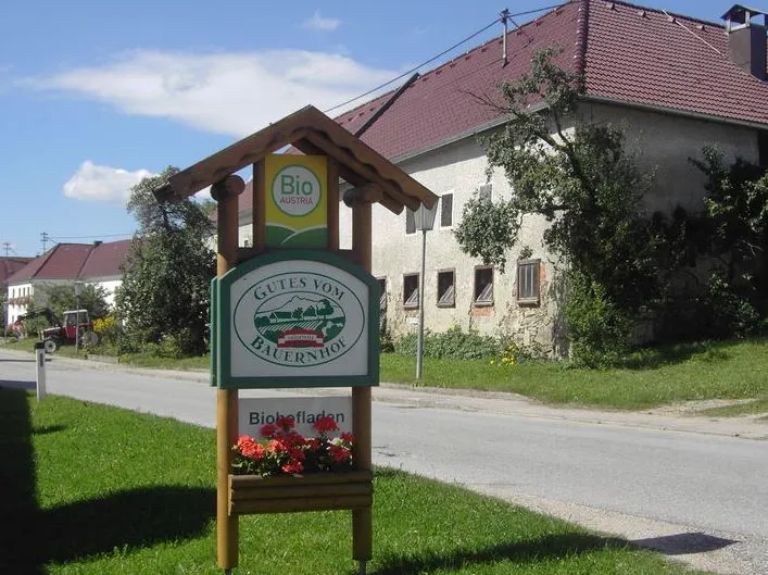 Auf dem Bild ist ein Bauernhof mit rotem Dach und grauer Fassade zu sehen; davor Wiese und ein Weg; davor das GvB Schild  mit Blumen