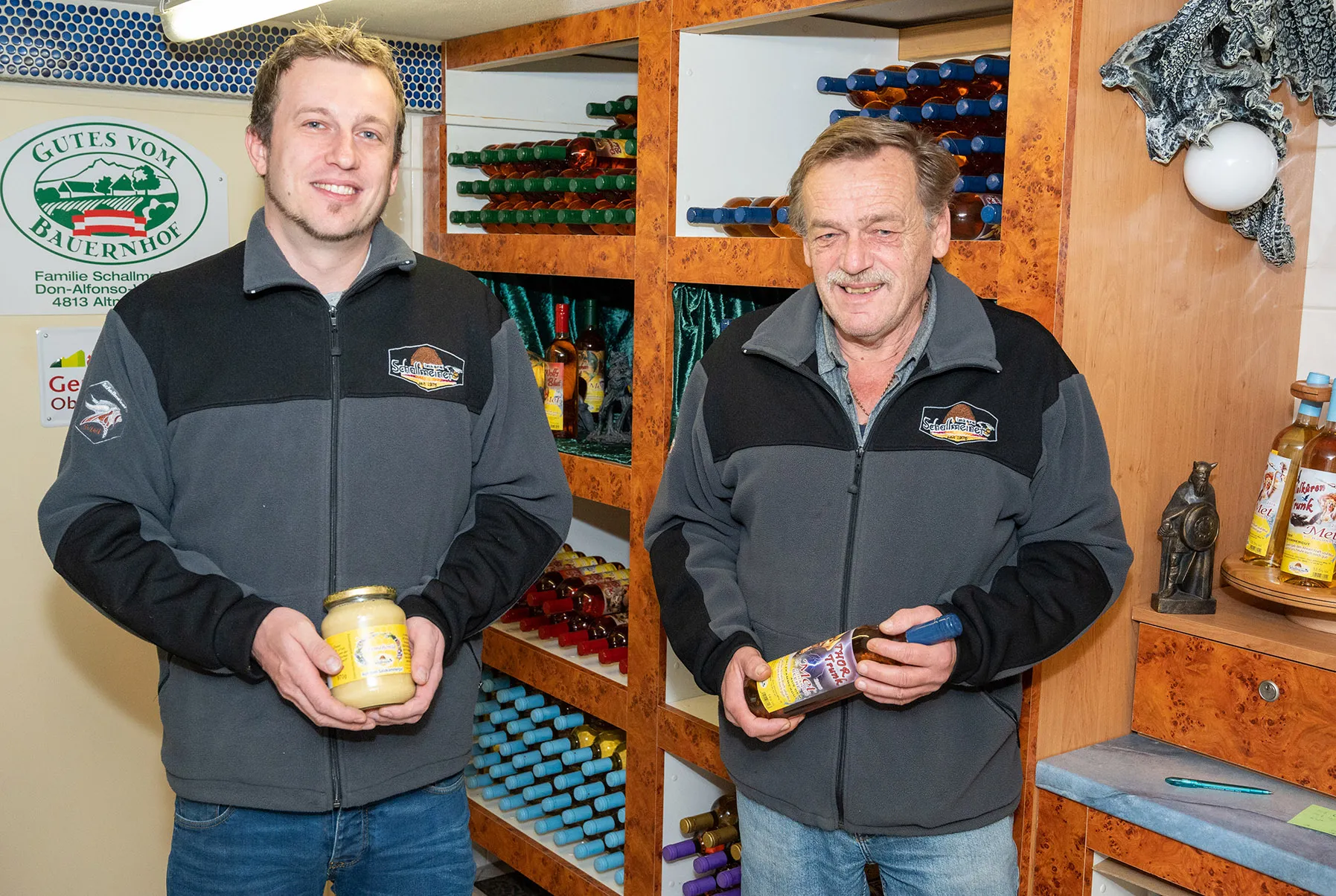 Herr Schallmeiner und sein Sohn stehen vor einem Regal mit Honigprodukten. Herr Schallmeiner jun. hält ein Glas Honig in den Händen, Herr Schallmeiner sen. eine Flasche 