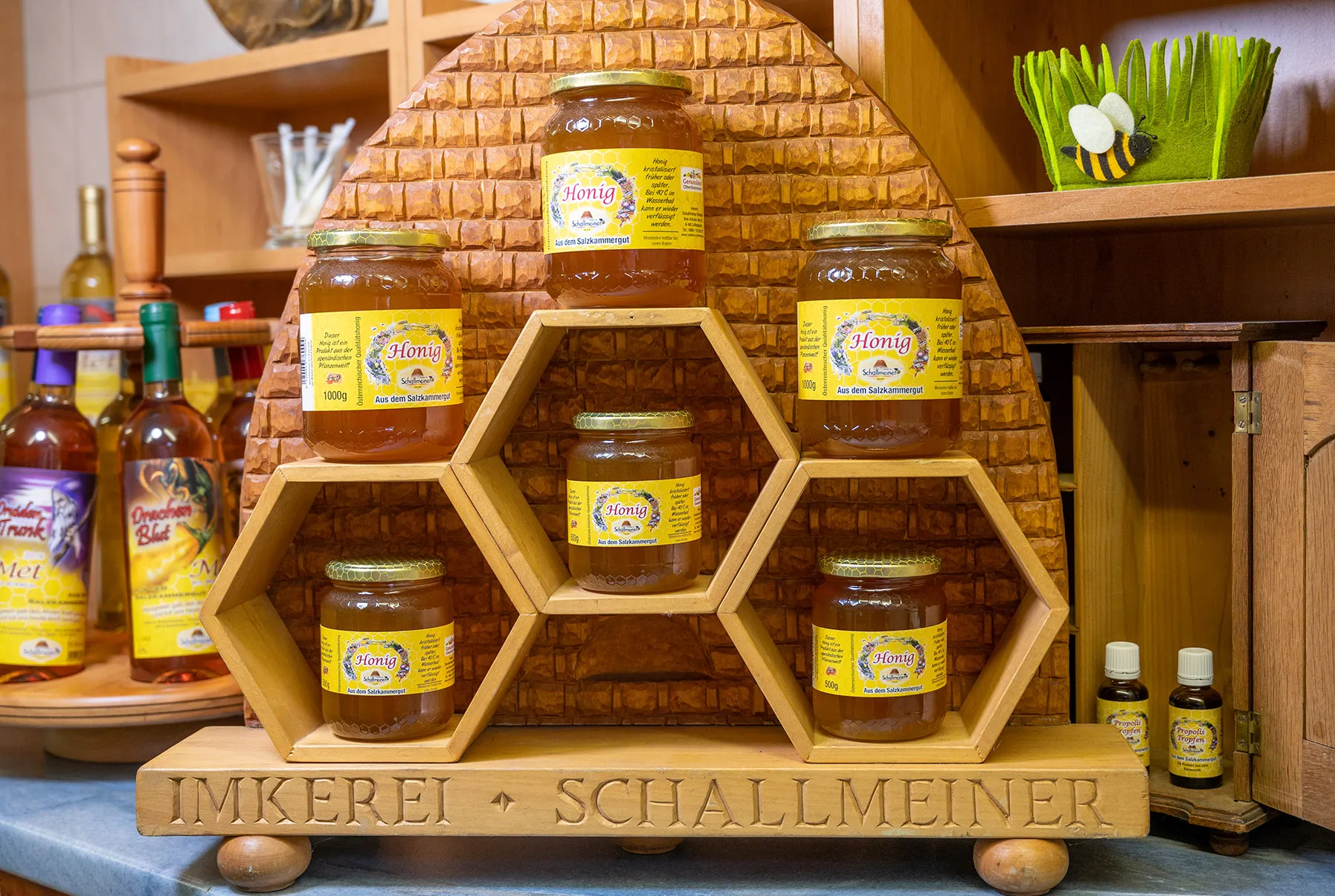 Auf dem Bild steht eine hölzerne Bienenwabe mit verschiedenen Gläsern mit Honig auf einem Tisch. Dahinter verschiedene Produkte vom Honig und Löffel zum Verkosten