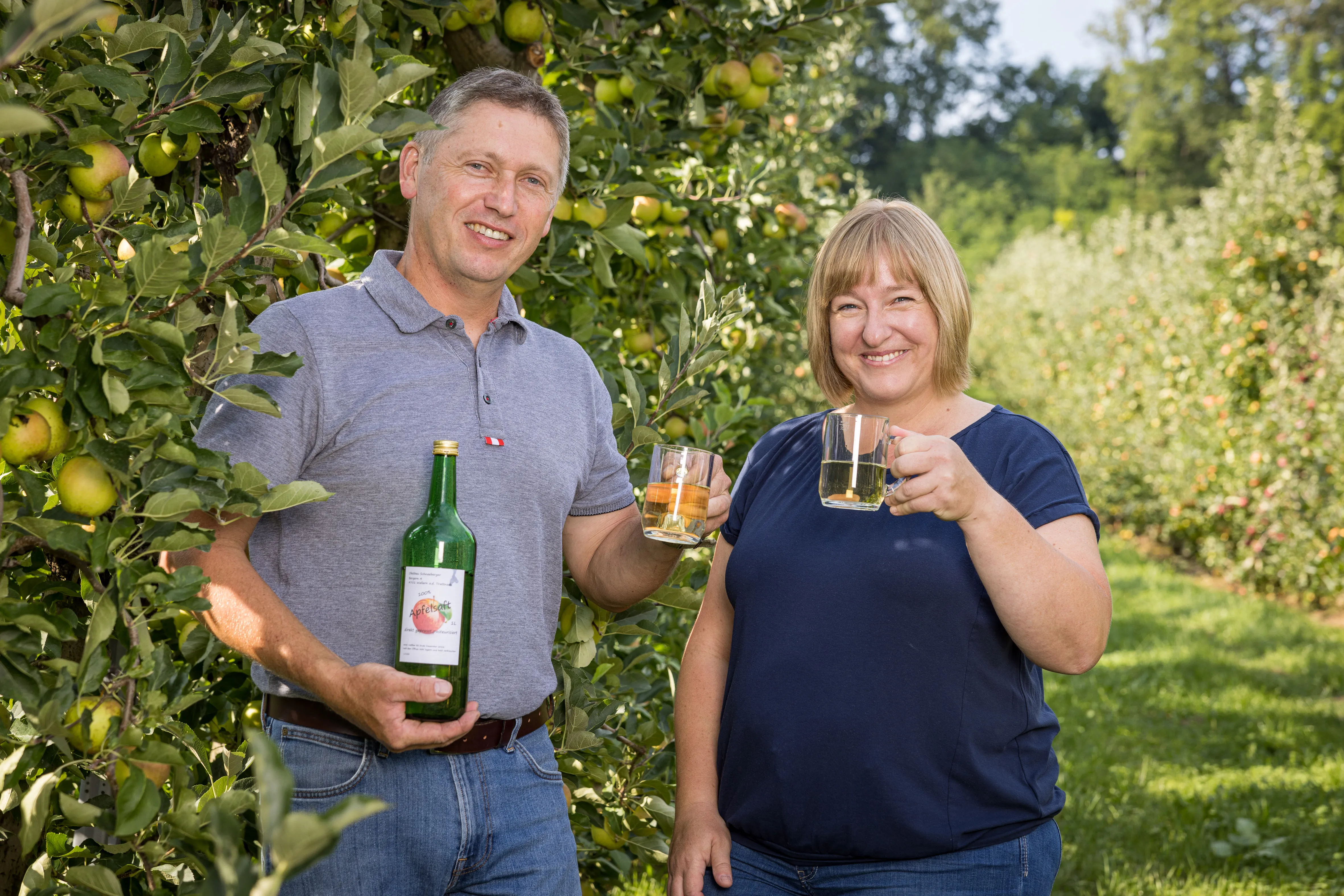 Ein Mann und eine Frau halten ein Glas bzw. eine Flasche in der Hand und stehen in einer Obstplantage vor einem Obstbaum