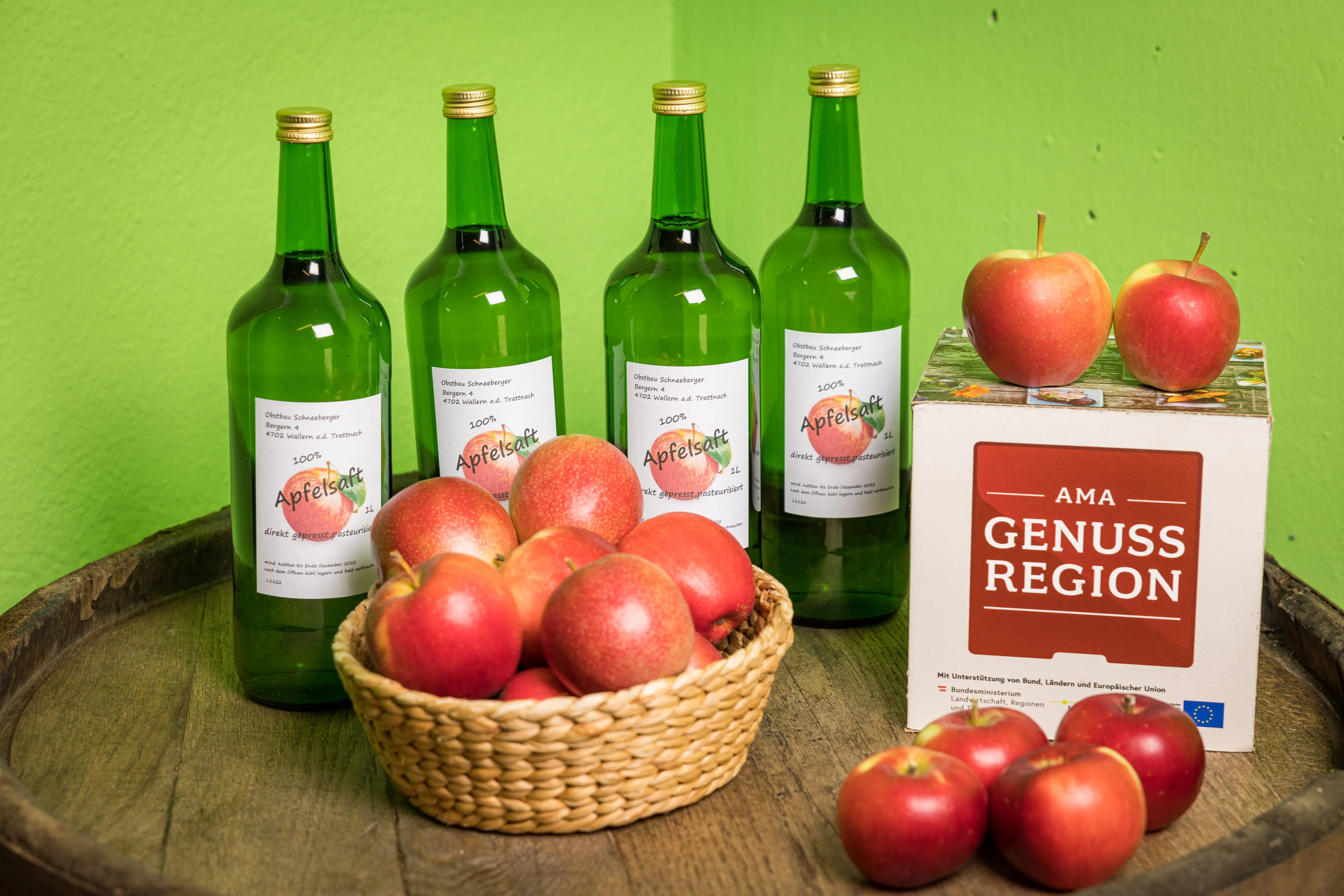 Auf einem Holzfass stehen vier Flaschen mit Apfelsaft, ein Korb mit roten Äpfeln, einige Äpfel lose und ein Kartonwürfel mit der Aufschrift AMA Genuss Region, grüne Wand im Hintergrund