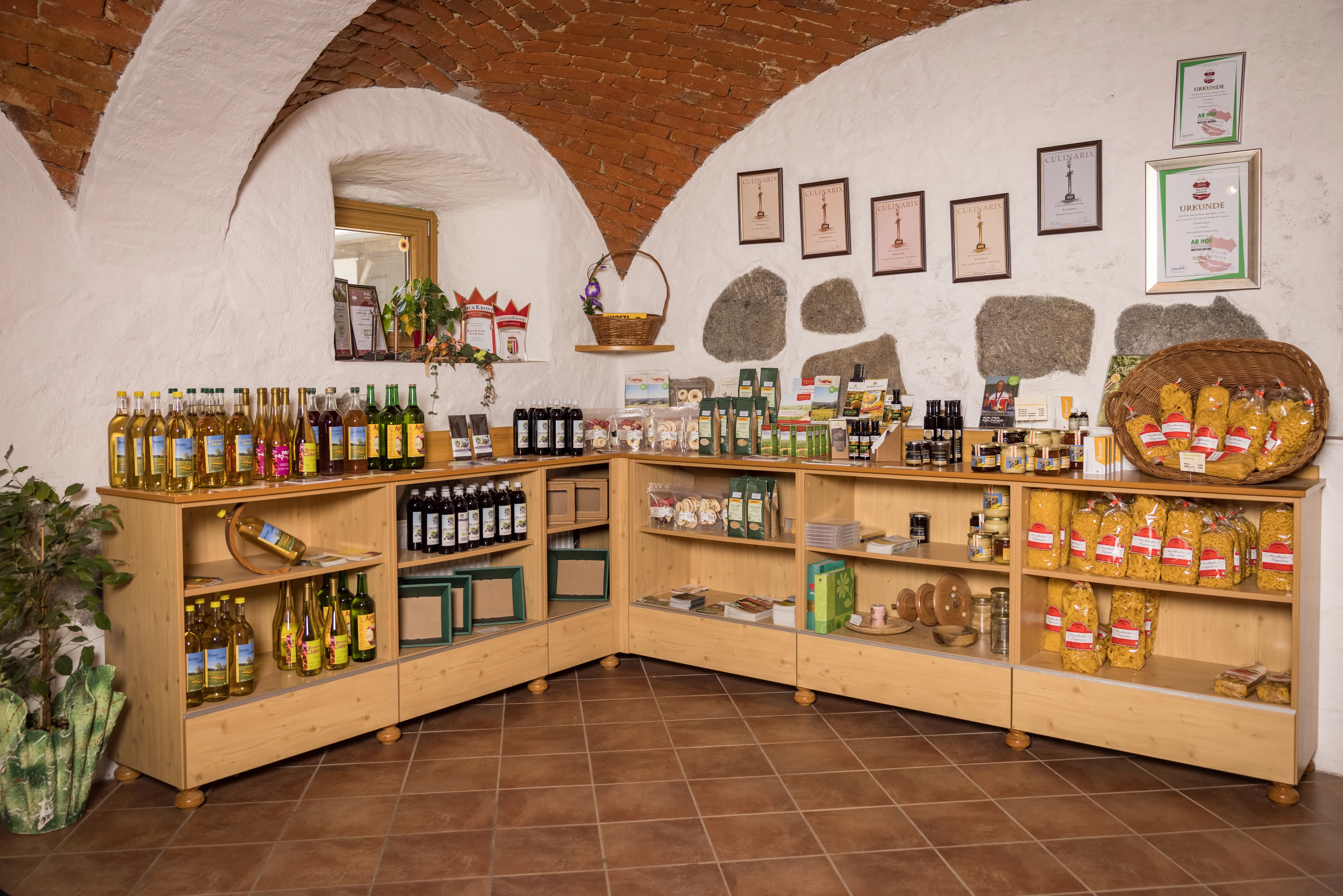 In einem Raum mit großem Gewölbe (weiß und braun gestrichen) stehen Holzregale an der Wand. Viele verschiedene Flaschen, Verpackungen, Packungen mit Nudeln, Gläser usw. stehen darin. An der Wand hängen mehrere Urkunden. Zur Deko Pflanzen.
