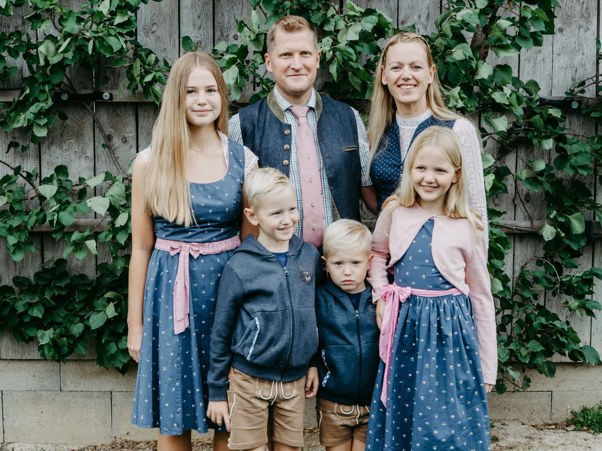 Ein Mann und eine Frau mit ihren vier Kindern stehen vor einem Holzgebäude, welches mit Efeu überwuchert ist.
