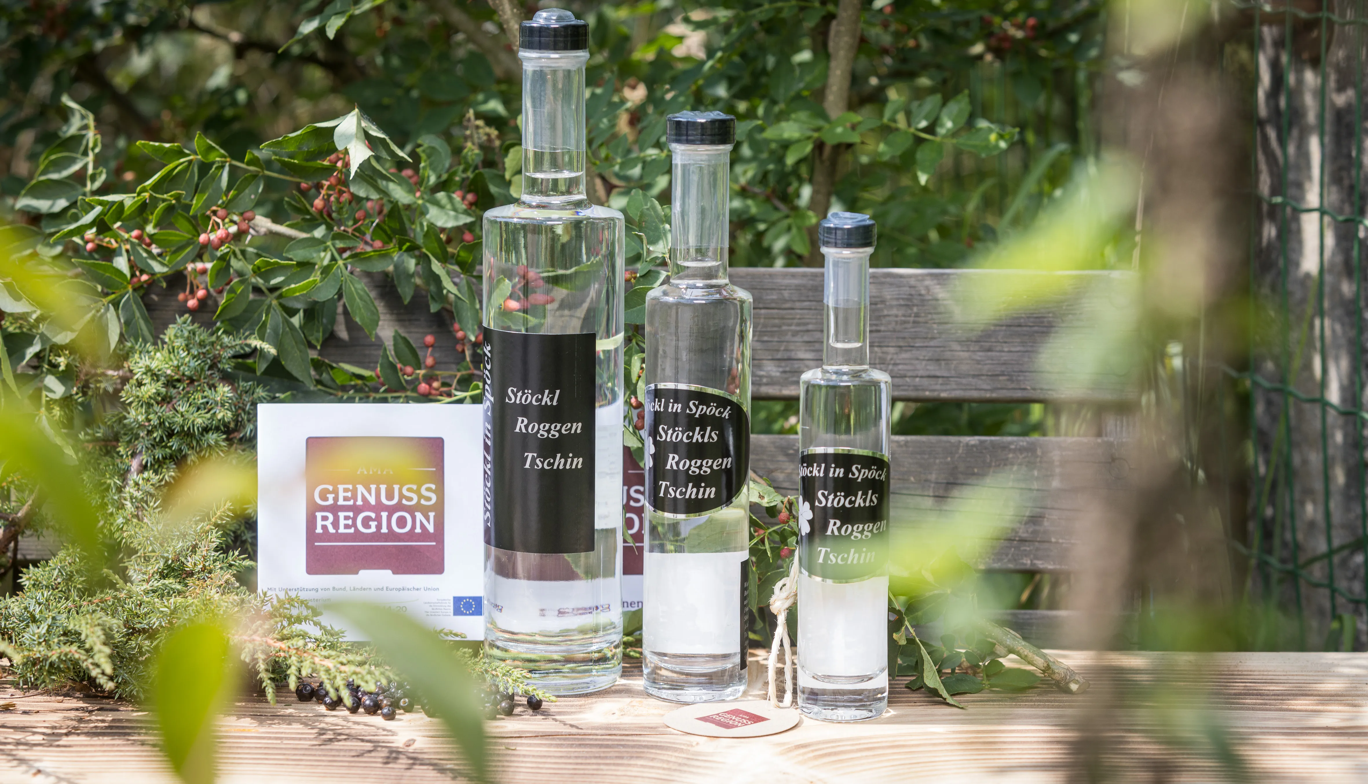 Auf einer Holzbank stehen mehrere Flaschen mit Tschin und ein Aufsteller Genuss Region, dahinter Blattwerk
