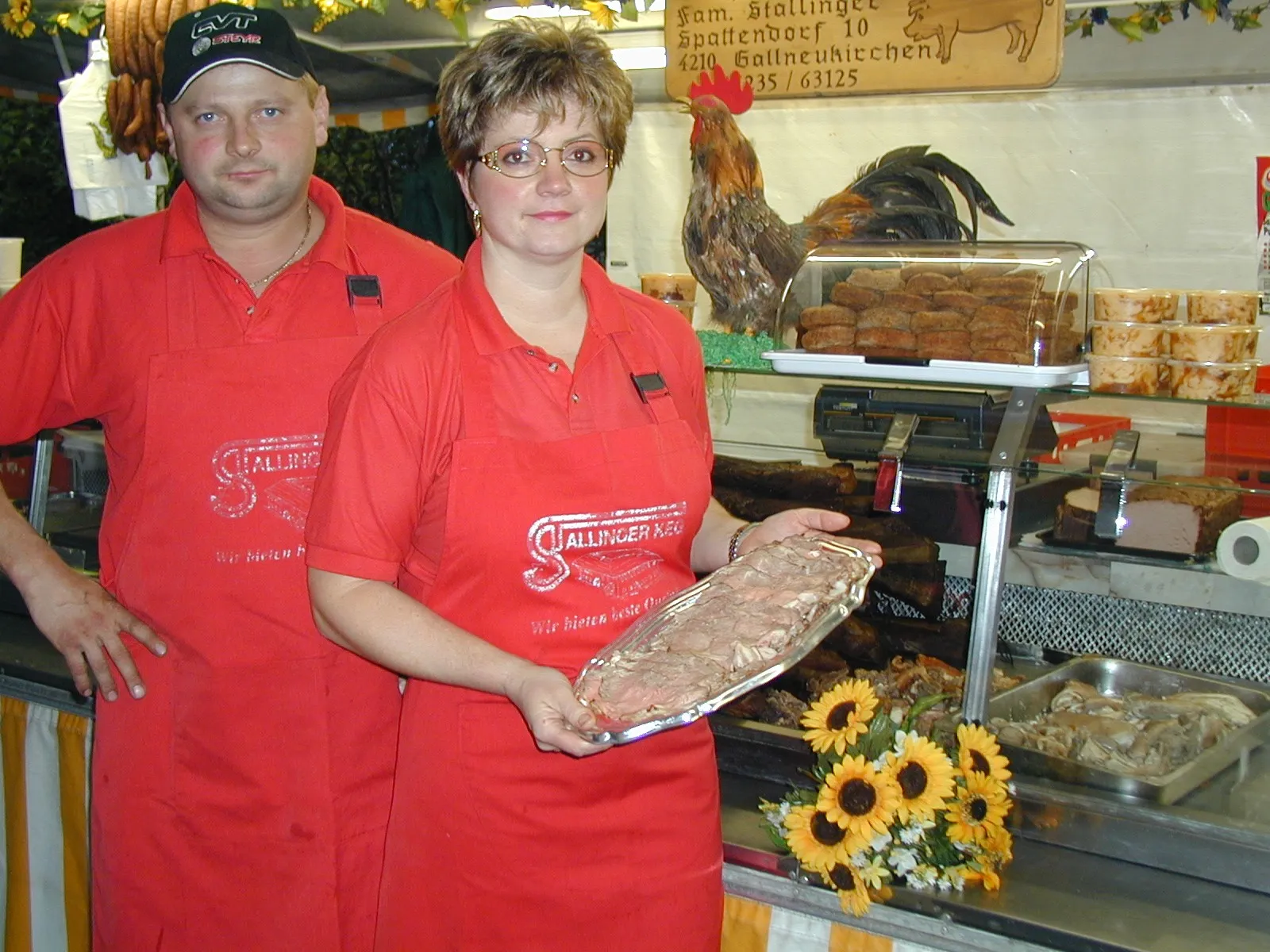 Ein Mann und eine Frau im roten Gewand und roter Schürze stehen vor einer Kühlvitrine; die Frau hält ein Brett mit aufgeschnittenem Speck in der Hand.