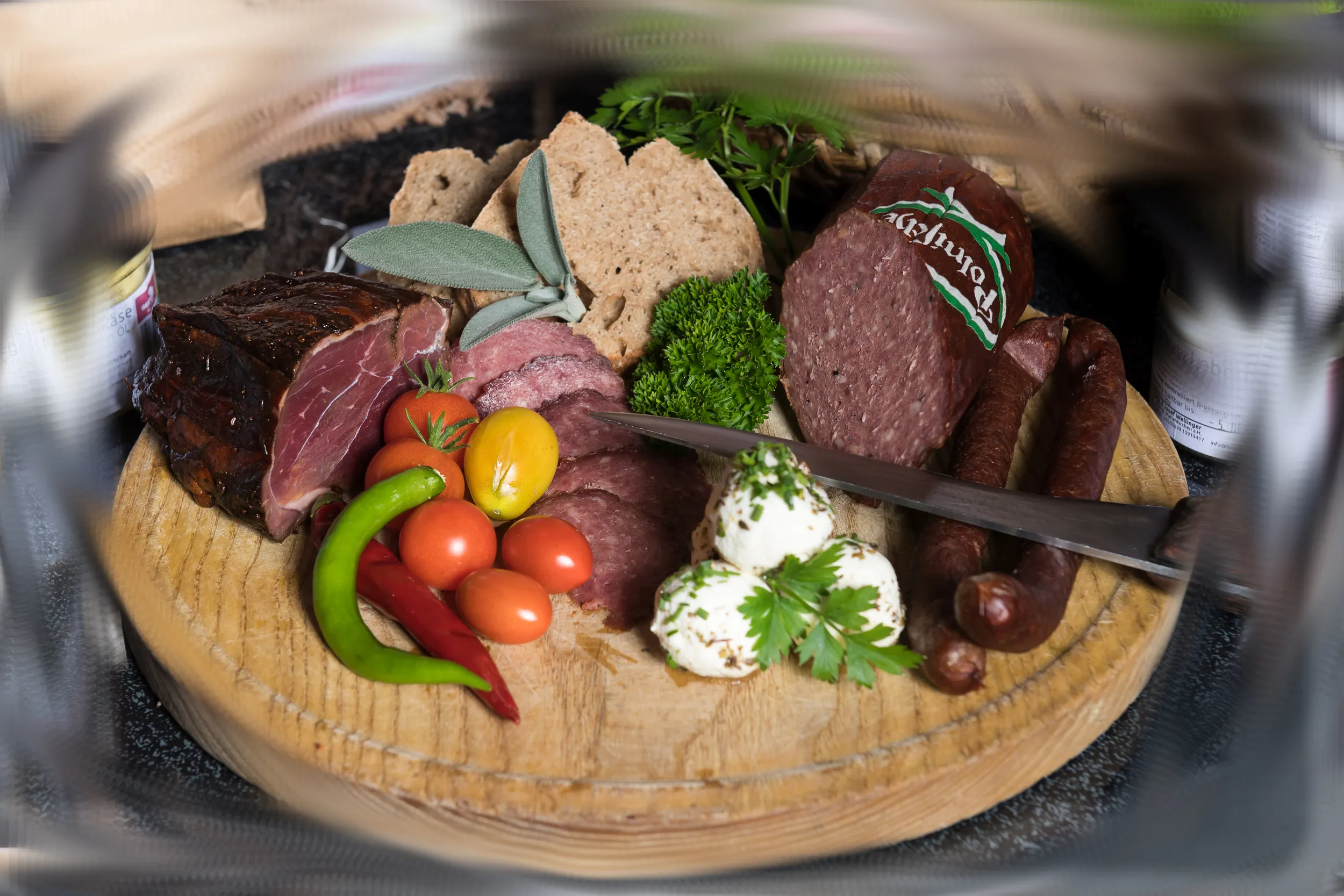 Auf einem Jausenbrett liegen eine Wurst, ein Stück Speck und andere Spezialitäten. Garniert mit Tomaten, Käse und Pfefferoni.