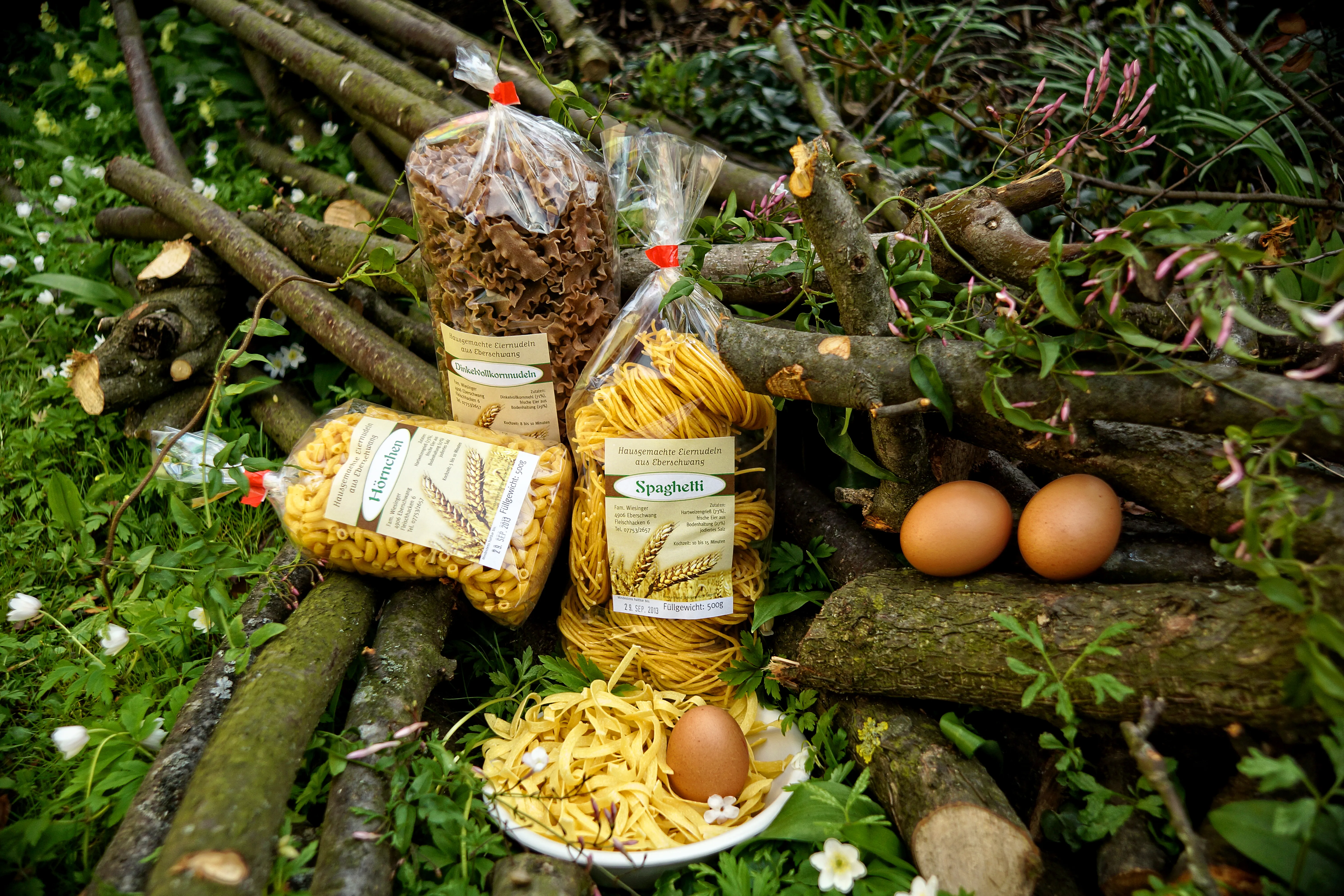 Auf einer Wiese, zwischen Totholz, liegen mehrere Packungen Nudeln, ein Teller mit Nudeln, und braune Eier.