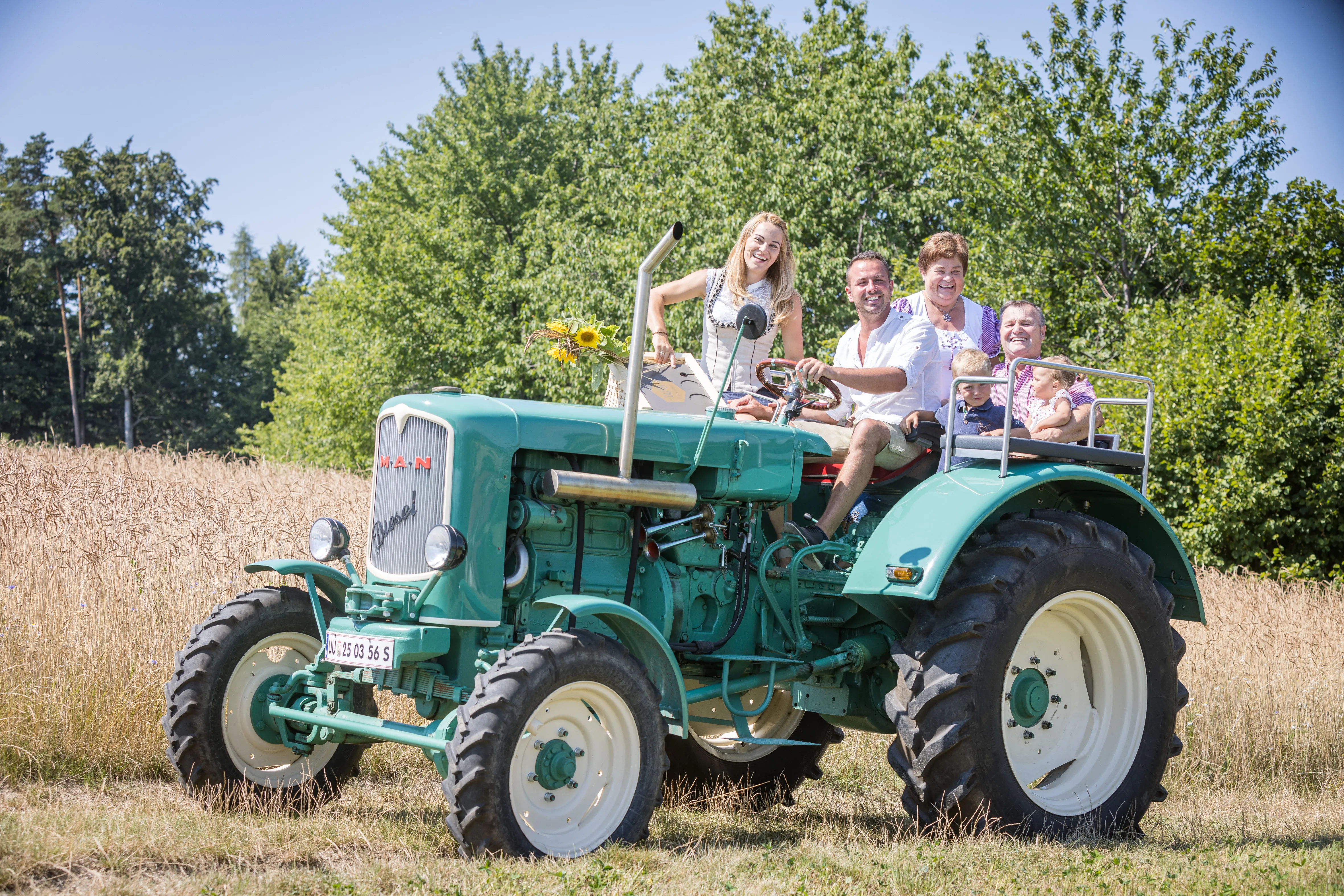 Auf einem grünen Traktor sitzen zwei Männer, zwei Frauen und zwei Kinder. Dahinter ein Feld, Bäume und Büsche und Himmel