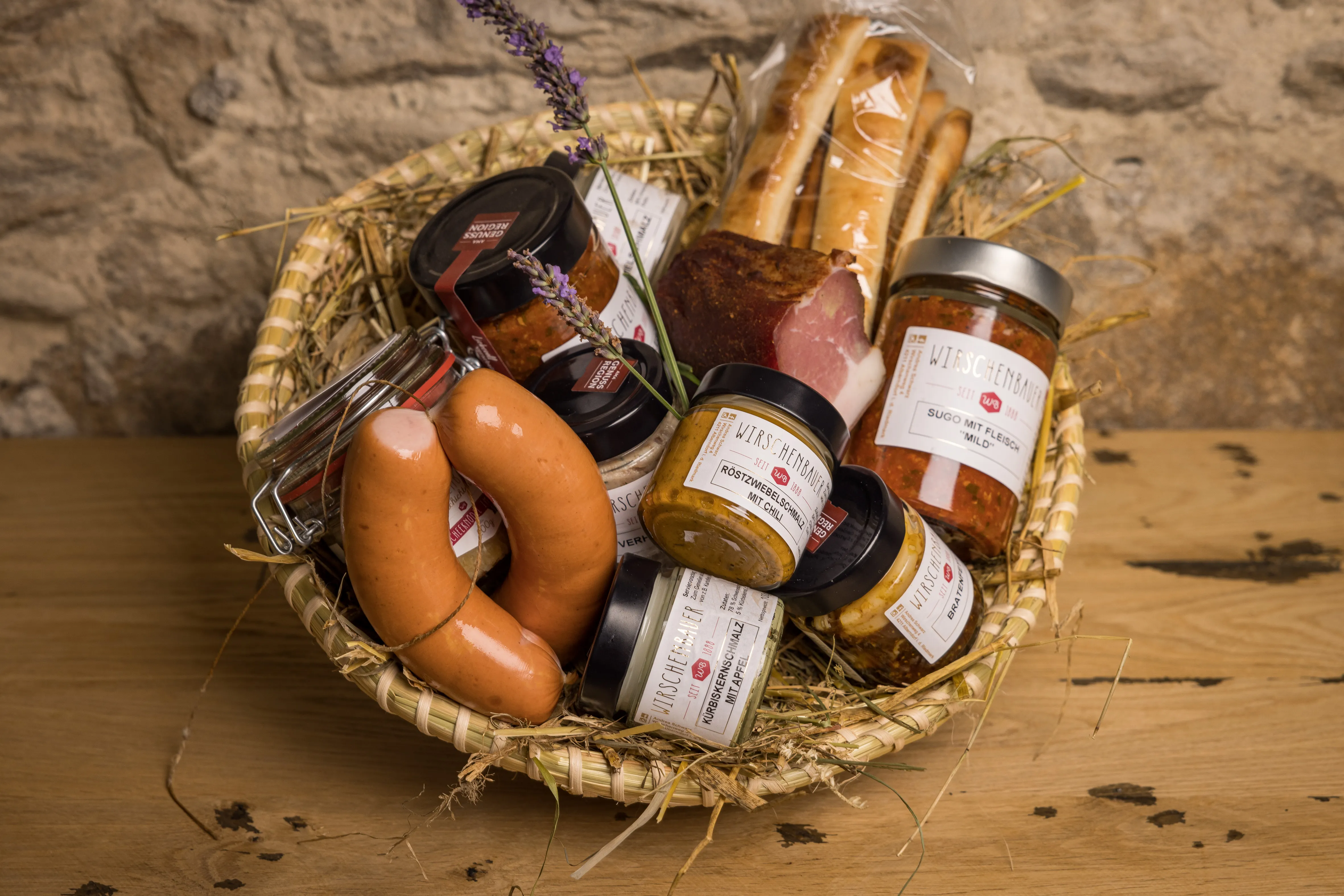 Auf einem Holztisch steht ein Korb mit Speck, Knacker, und verschieden großen Gläsern mit Fertigprodukten.