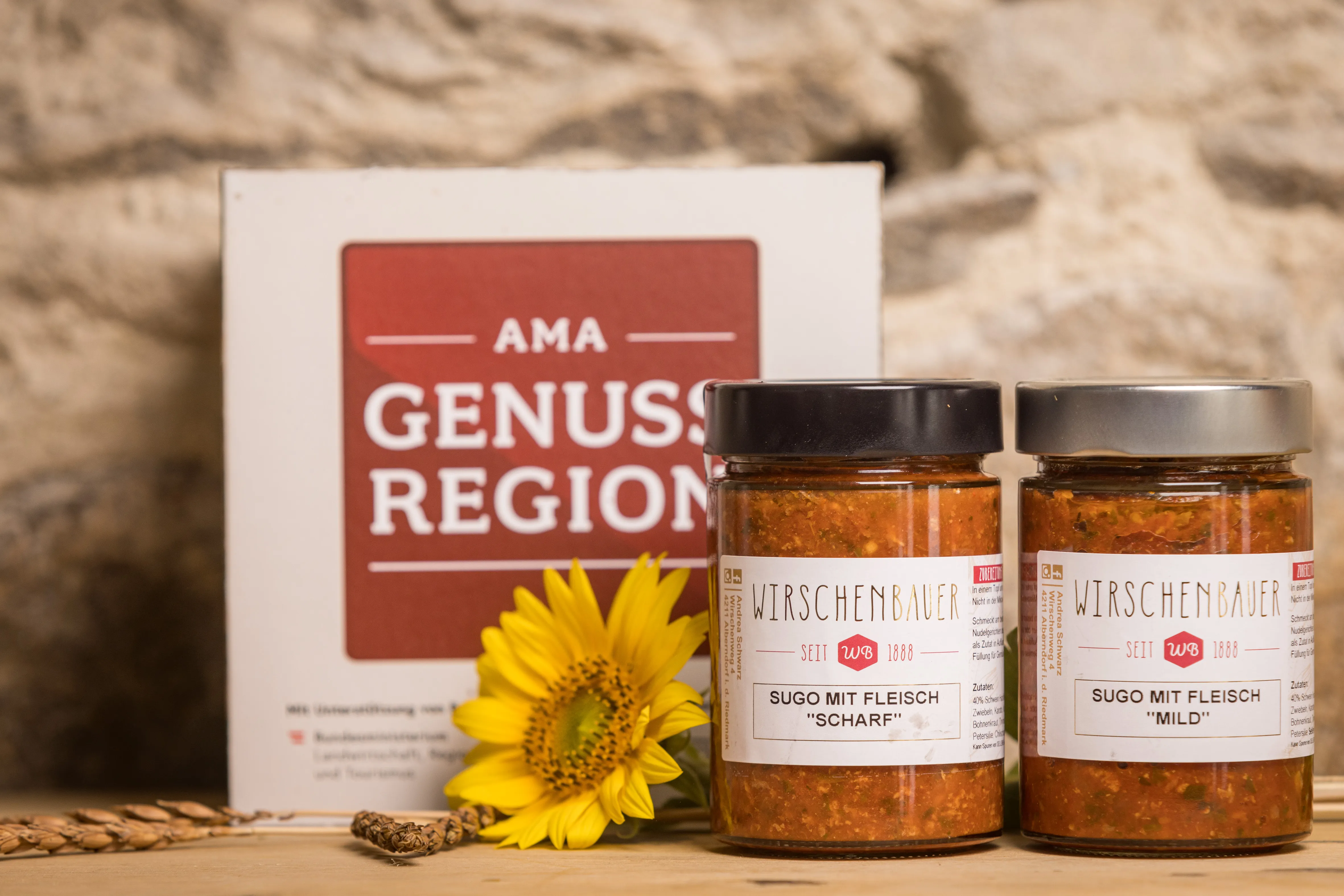 Auf einem Holztisch stehen zwei Gläser mit Fleischsugo, daneben eine Blüte einer Sonnenblume, dahinter Steinwand mit einem Aufsteller mit AMA Genuss Region Aufschrift