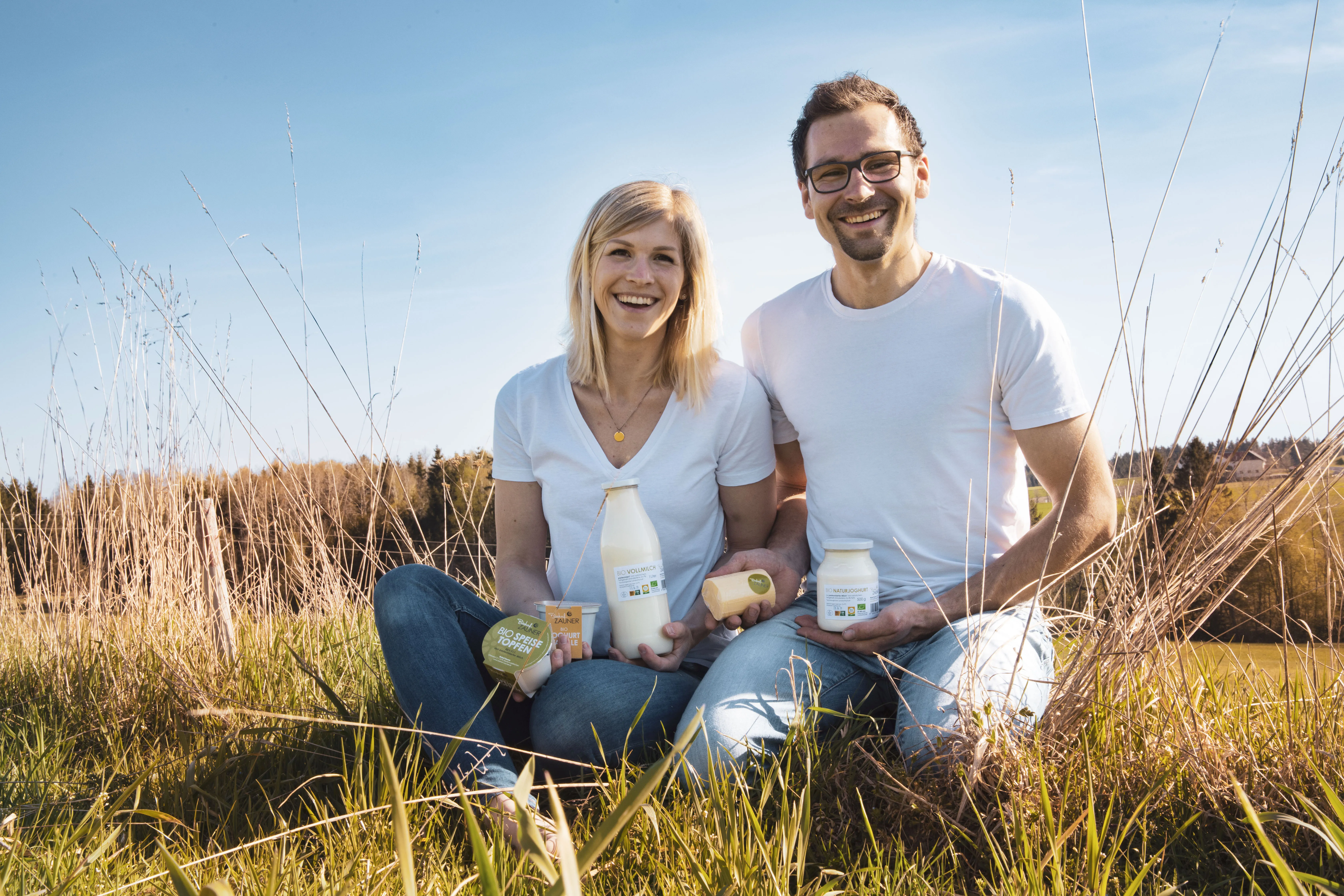 Ein Mann und eine Frau im weißen T-Shirt sitzen bzw. knien auf einem Feld, dahinter blauer Himmel