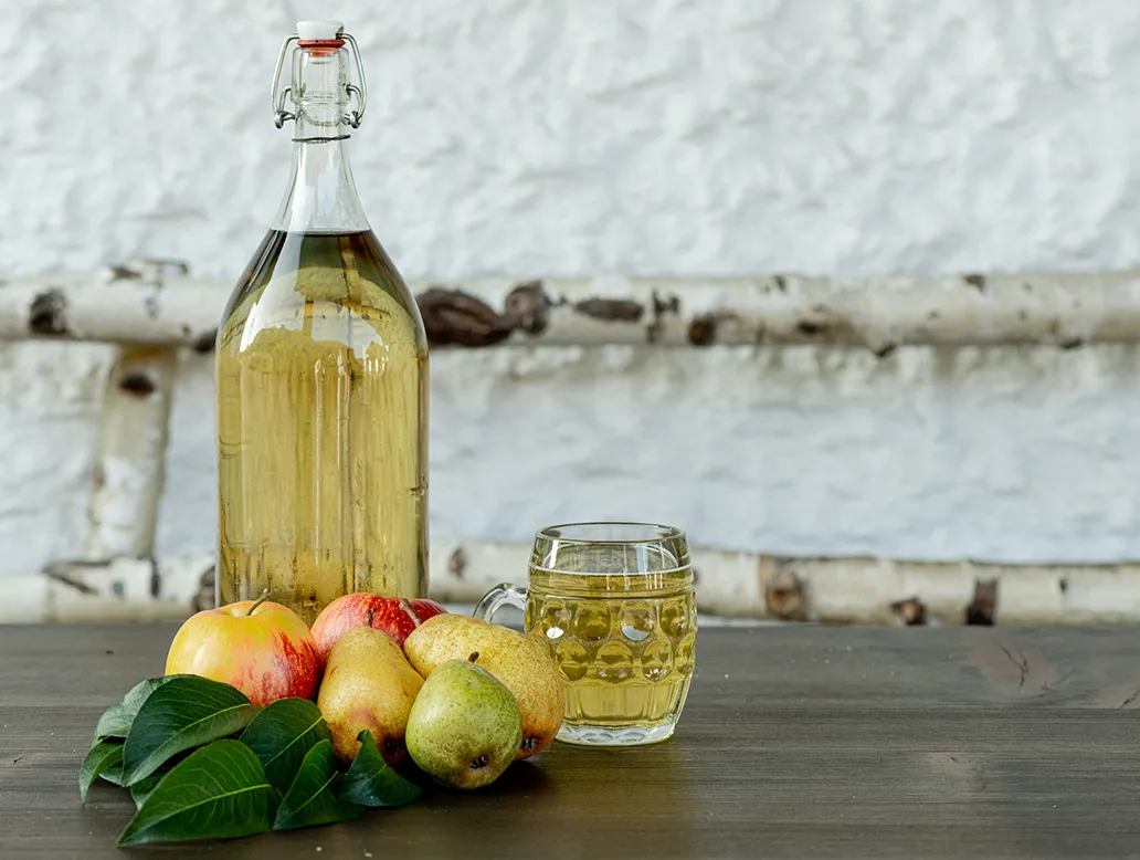Große Glasflasche und Glas mit hellem Getränk, davor Äpfel, Birnen und grüne Blätter auf Holztisch.