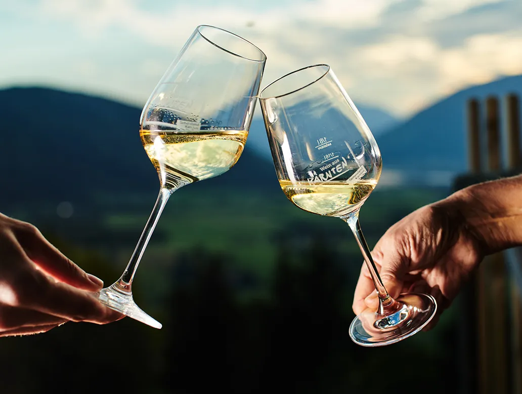 Zwei Hände stoßen mit Weingläsern mit hellem Wein vor einer Berglandschaft an.