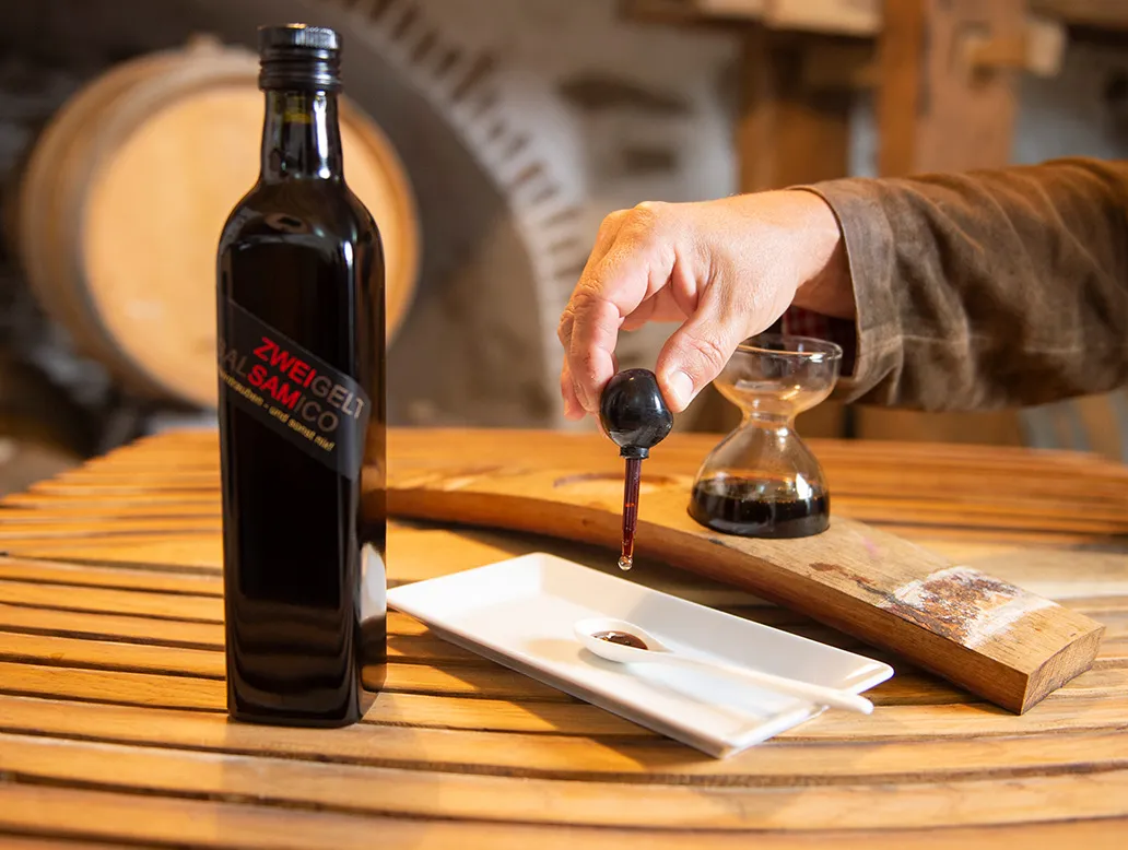 Hand tropft dunkle Flüssigkeit aus einer kleinen Flasche auf eine weiße rechteckige Schale, daneben steht eine dunkle Flasche mit rotem Etikett 'ZWEIGELT BALSAMICO'.