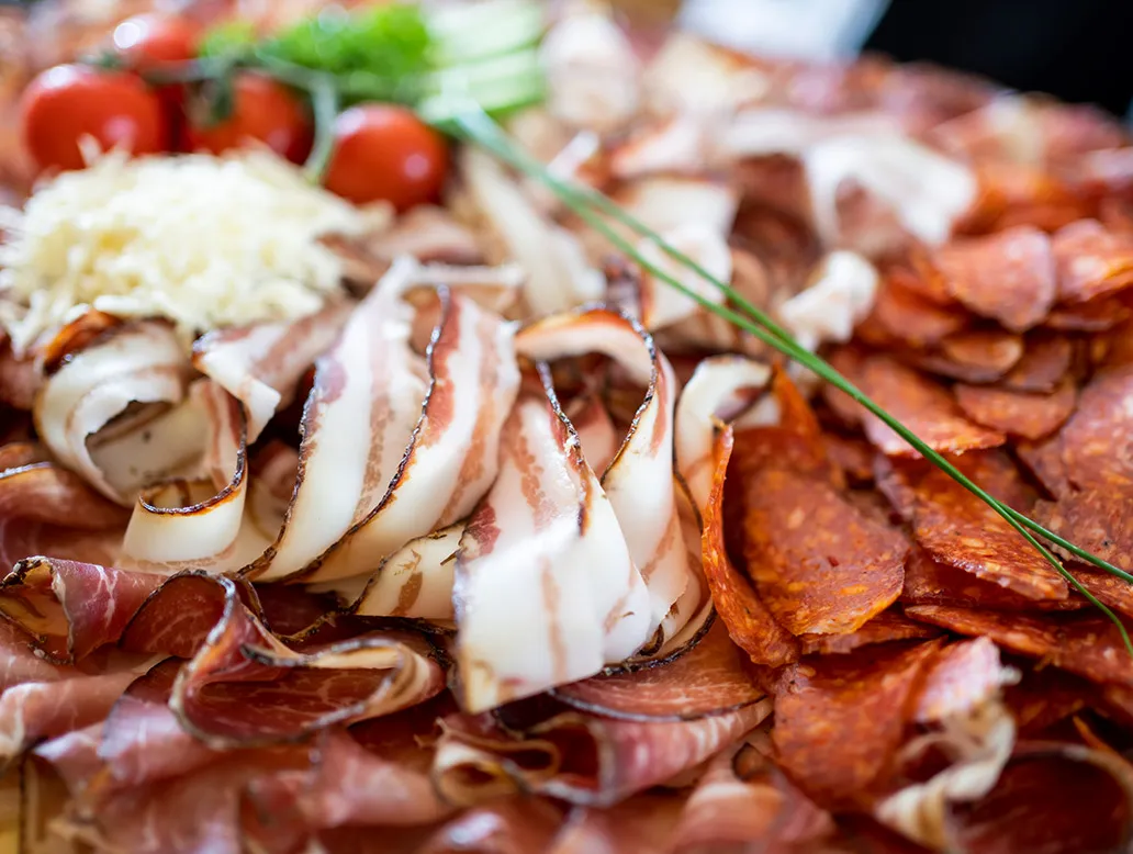 Nahaufnahme von dünn geschnittenem Speck und Paprikasalami auf einem Holzbrett, im Hintergrund Tomaten und Kräuter.