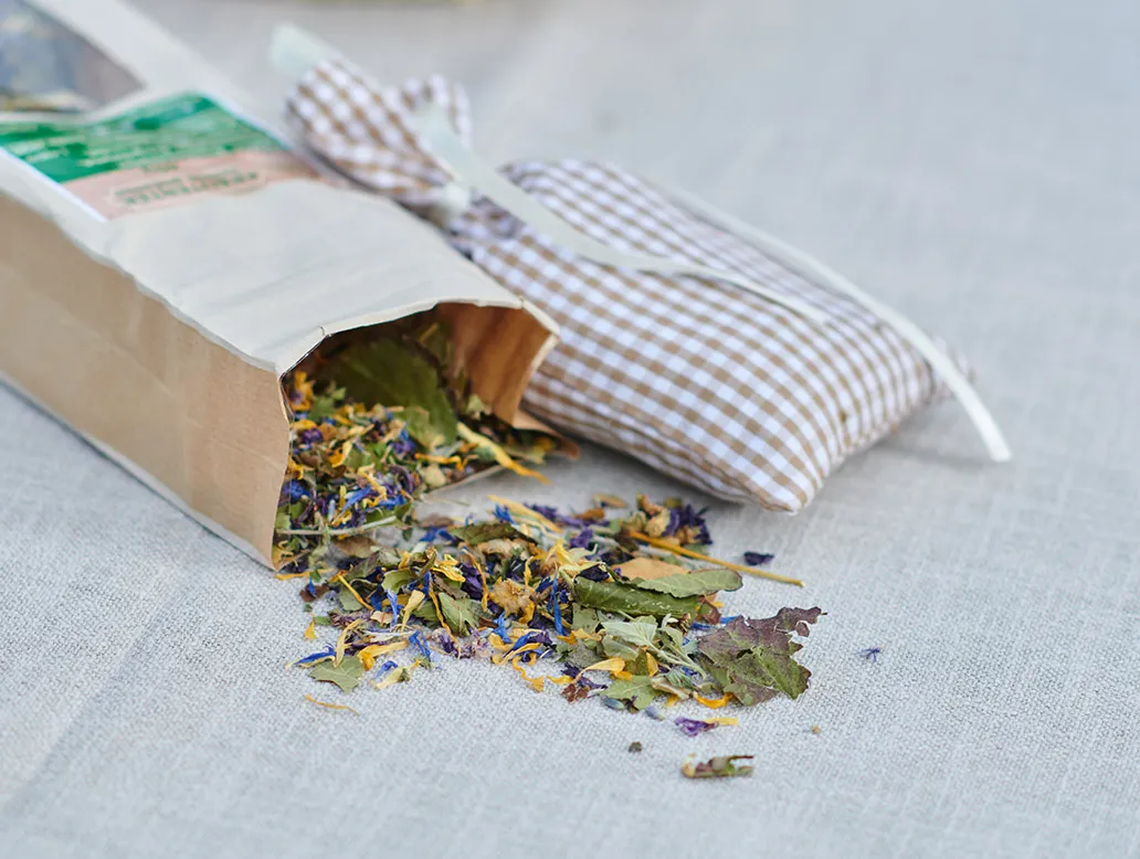 Braunes Papiertütchen mit getrockneten Kräutern und Blüten, daneben kariertes Stoffbeutelchen auf hellem Stoff.