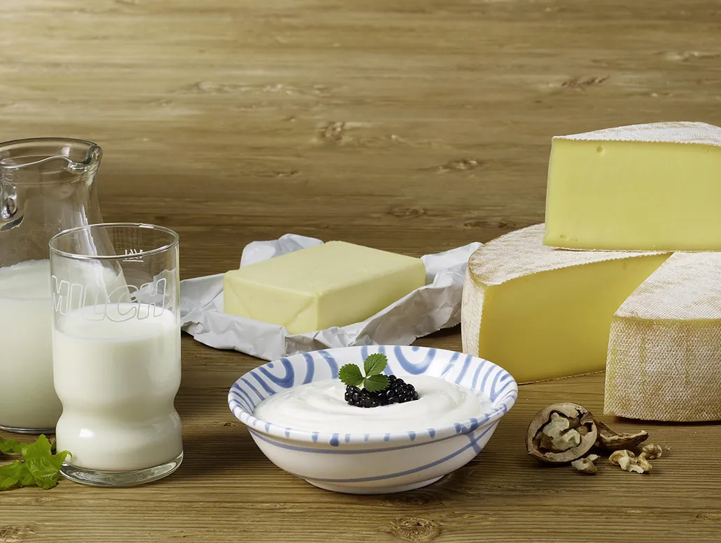 Großer runder Käse mit einem Stück herausgeschnitten, Butter in Folie, Glas und Karaffe mit Milch, Schale mit Quark und Beeren, Walnüsse und Trauben auf Holzuntergrund.