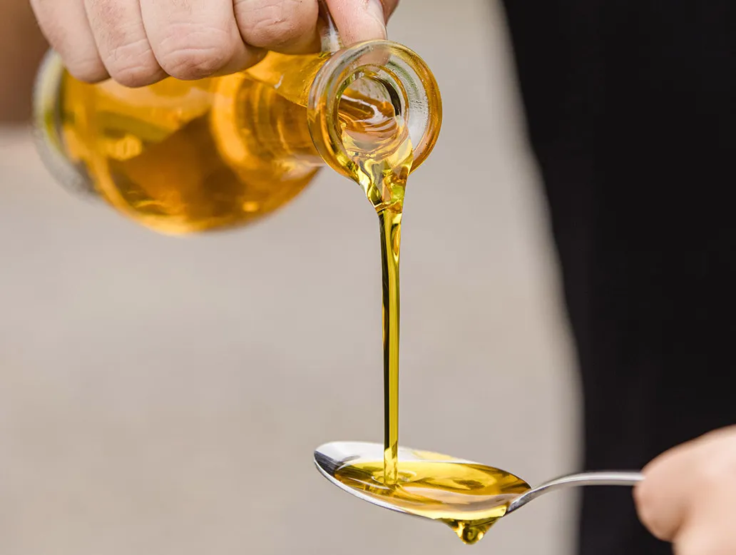 Hand gießt goldgelbes Öl aus einer Glasflasche in einen Metalllöffel.