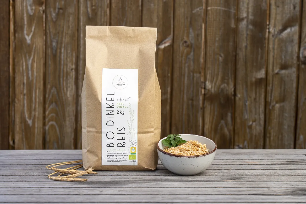 Dinkelreis in Verpackung auf einem Holztisch vor einer Holzwand, rechts daneben fertig gekochter Dinkelreis in Schale