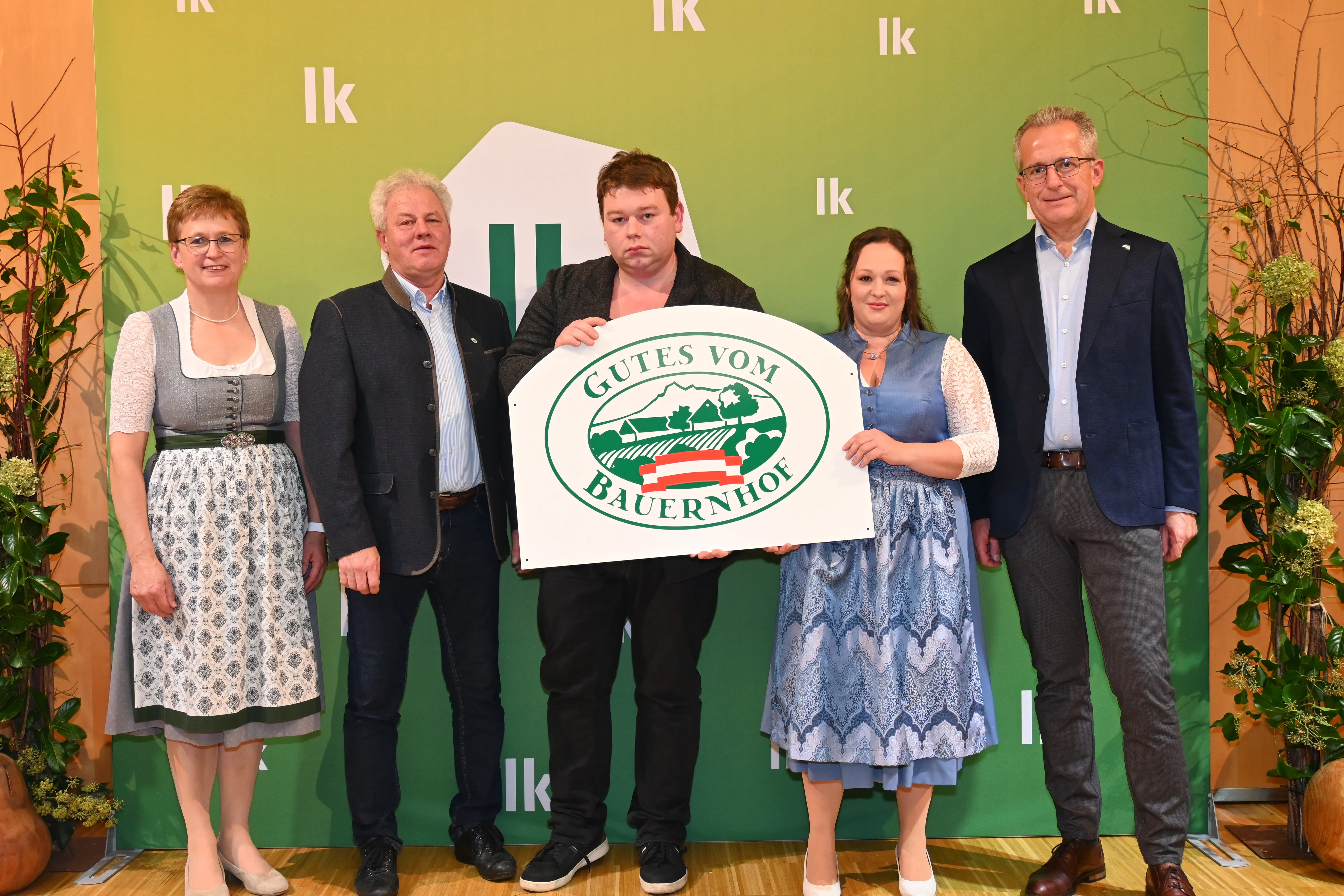 Auf einer Bühne mit grünem Hintergrund stehen Lea Marie und Roland Lamberg mit einer Hoftafel in den Händen. Daneben zwei weitere Männer und eine Frau, alle in Tracht.