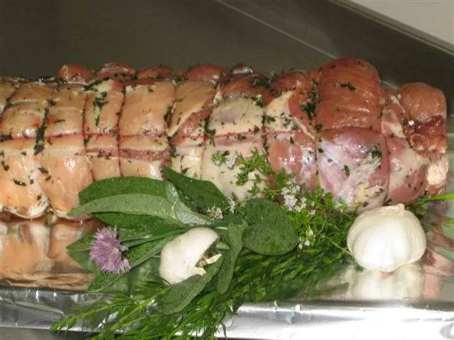 man sieht einen rohen Kalbsrollbraten gewürzt mit Kräuter, daneben Knoblauch und Kräuter