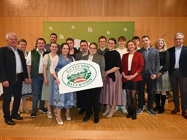 Gruppe von Menschen in traditioneller und moderner Kleidung steht vor grünem Hintergrund mit weißem Schriftzug 'lk'. Eine Person hält ein Schild mit grün-rotem Logo und Schriftzug 'Gutes vom Bauernhof'.