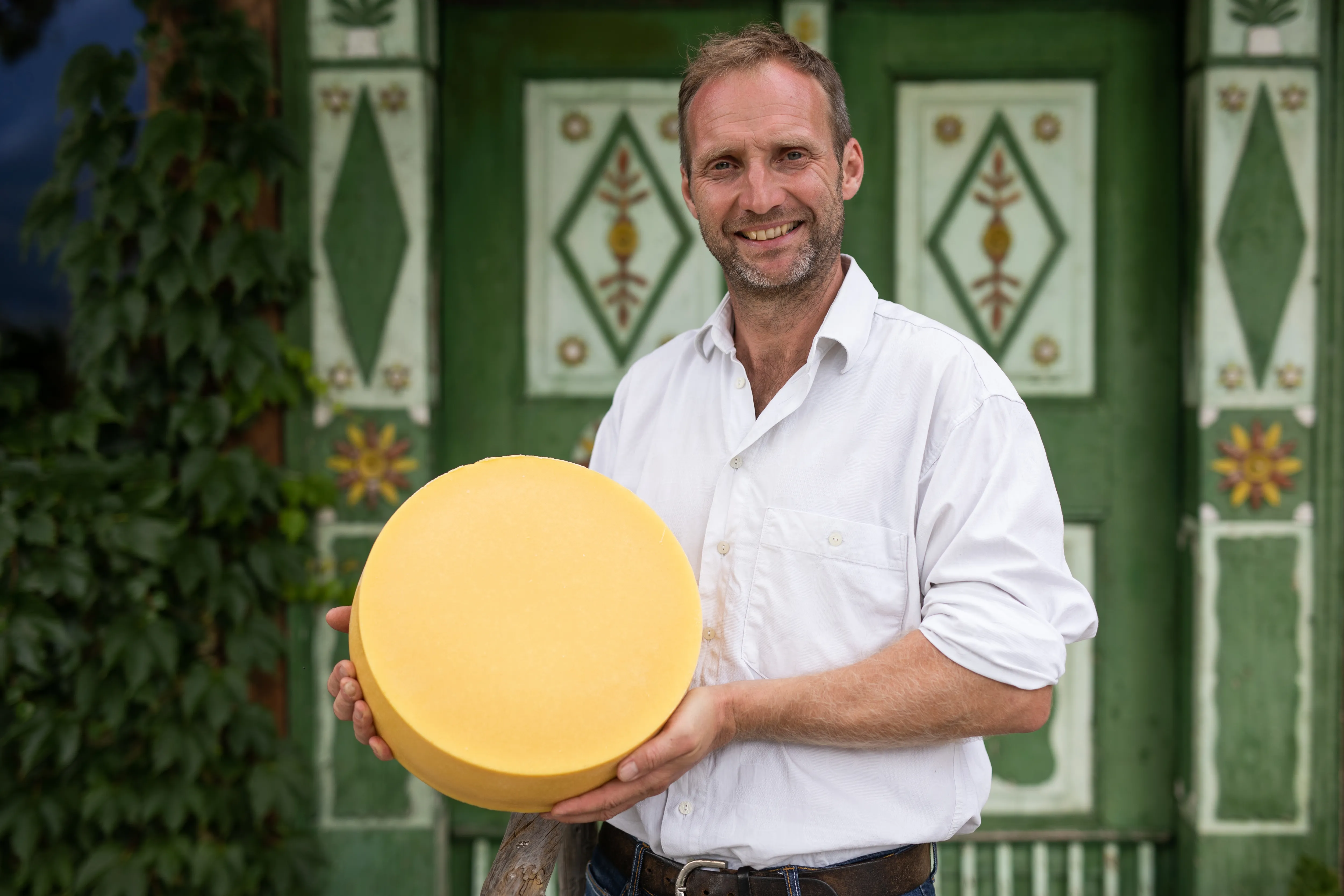 Niki Rettennacher vom Fürstenhof mit einem Käselaib stehend vor einer bunten alten Holztür