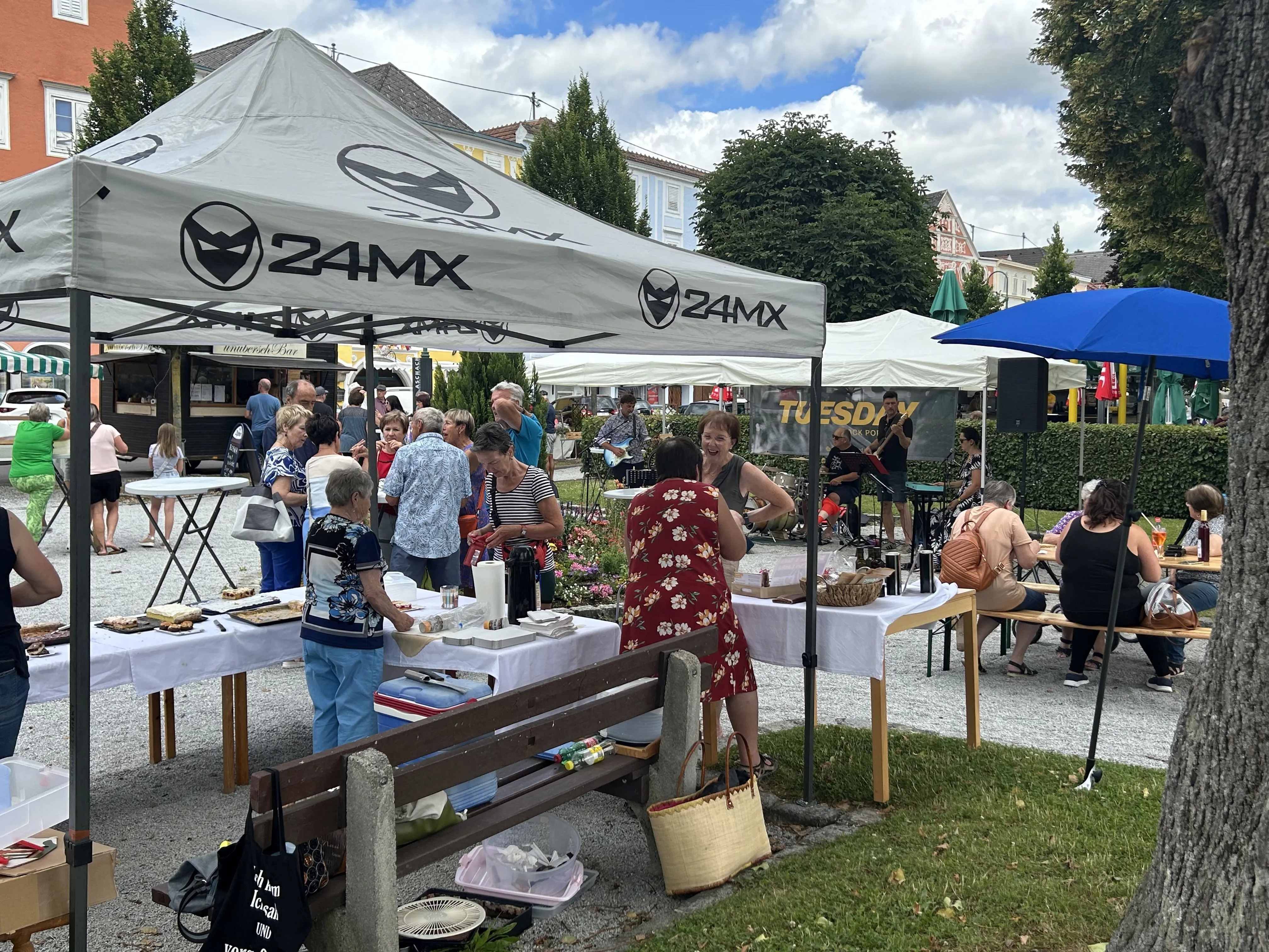 Auf dem Bild sieht man mehrere Marktstandl mit verschiedenen Personen. Einige Personen sitzen auf einer Bank. Im Hintergrund Häuserfronten, grüne Bäume und blauer Himmel mit Wolken