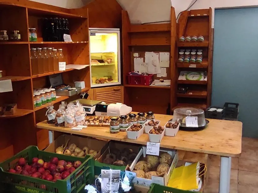 In einem Raum mit Gewölbe stehen an der Wand mehrere Regale und ein Kühlschrank. In den Regalen verschiedene Flaschen und Produkte; davor ein Tisch mit einer Waage und mehreren Produkten. Davor noch Kisten mit Gemüse und Obst. 