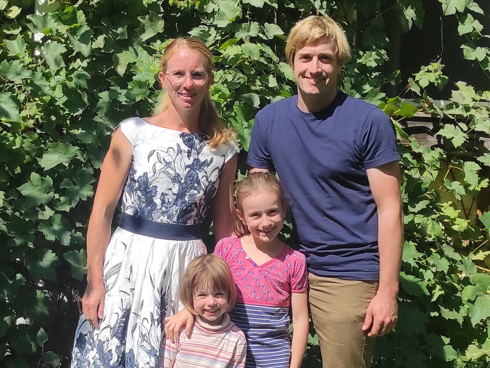 Herr und Frau Blümel stehen mit ihren beiden Mädchen vor einer grünen Hecke. Das größere Kind hat die Hand um die Schultern des kleineren Kindes gelegt. 