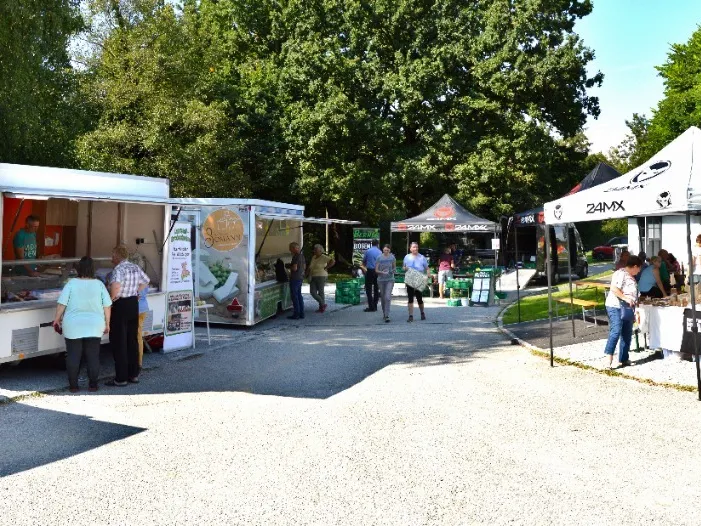 Verschiedene Stände auf einem Bauernmarkt, Personen und grüne Bäume im Hintergrund