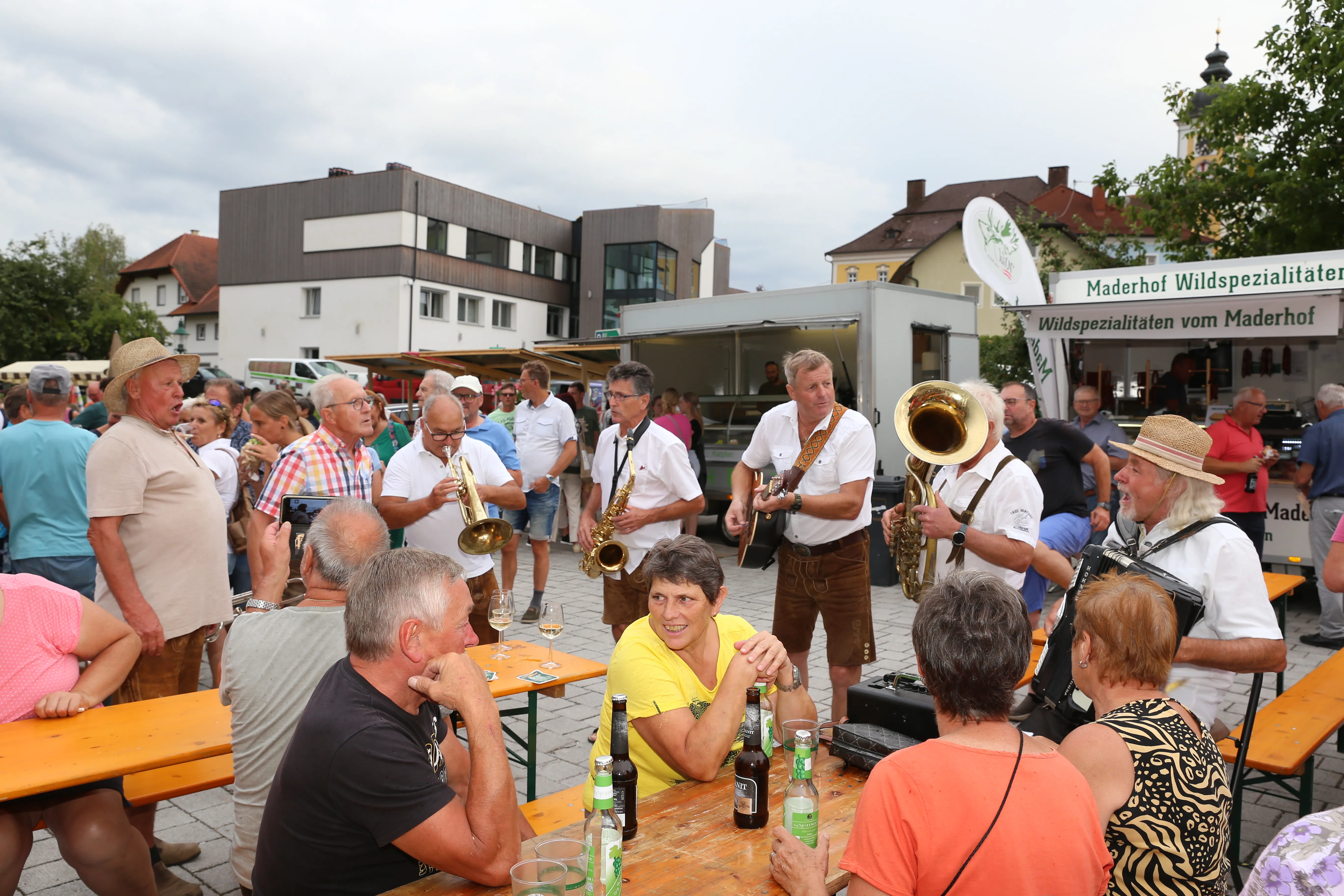 Auf dem Foto sieht man eine Musikgruppe auf einem Marktplatz, dahinter mehrere Stände, davor Tische und Bänke mit einigen Leuten.