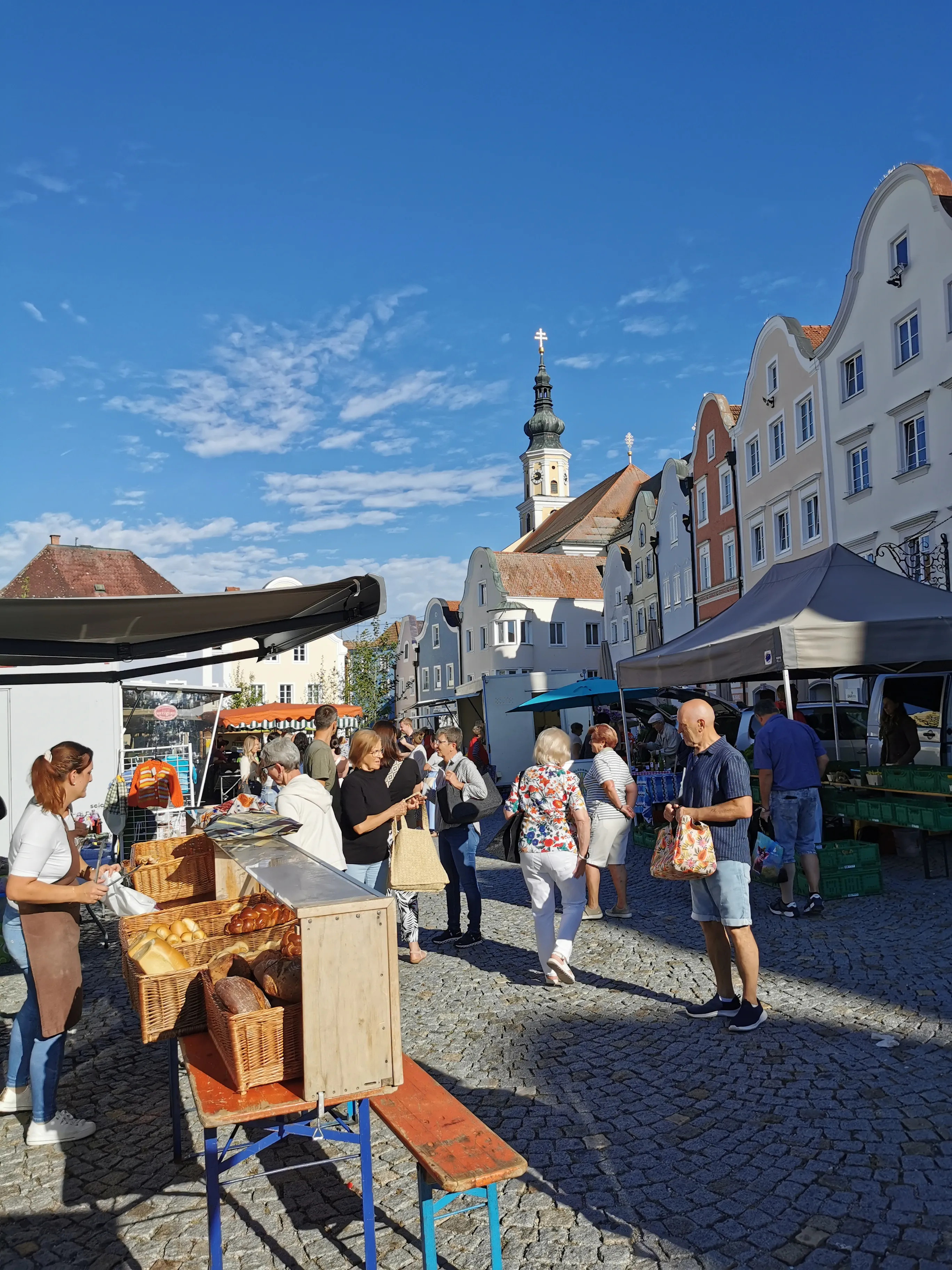 Personen und Marktstände, im Hintergrund eine Kirche und Häuserfassaden, dahinter blauer Himmel
