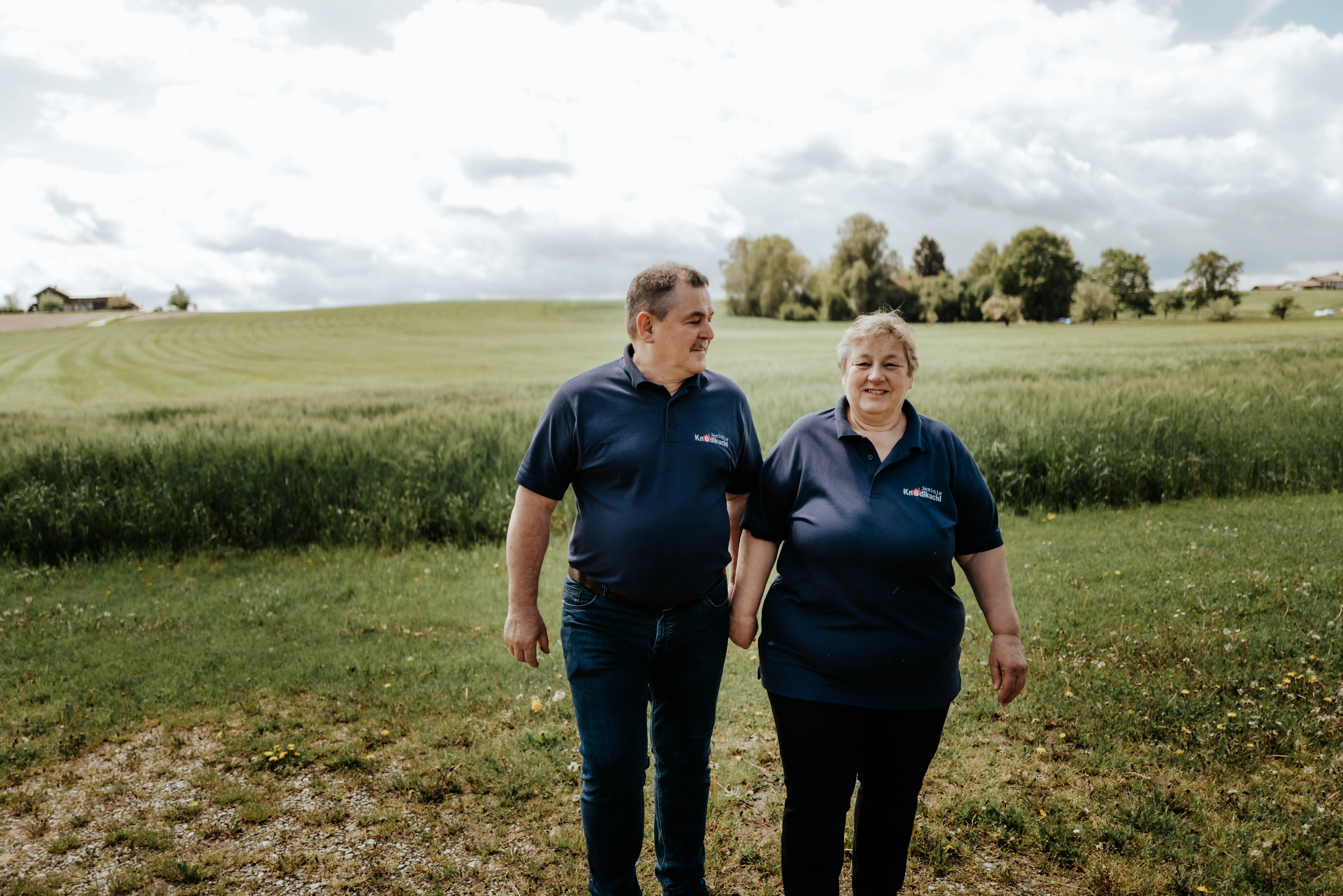 Herr und Frau Jenichl, beide dunkel gekleidet, stehen auf einer Wiese; im Hintergrund grüne Felder, Bäume und mit Wolken bedeckter Himmel; ganz hinten ein Haus