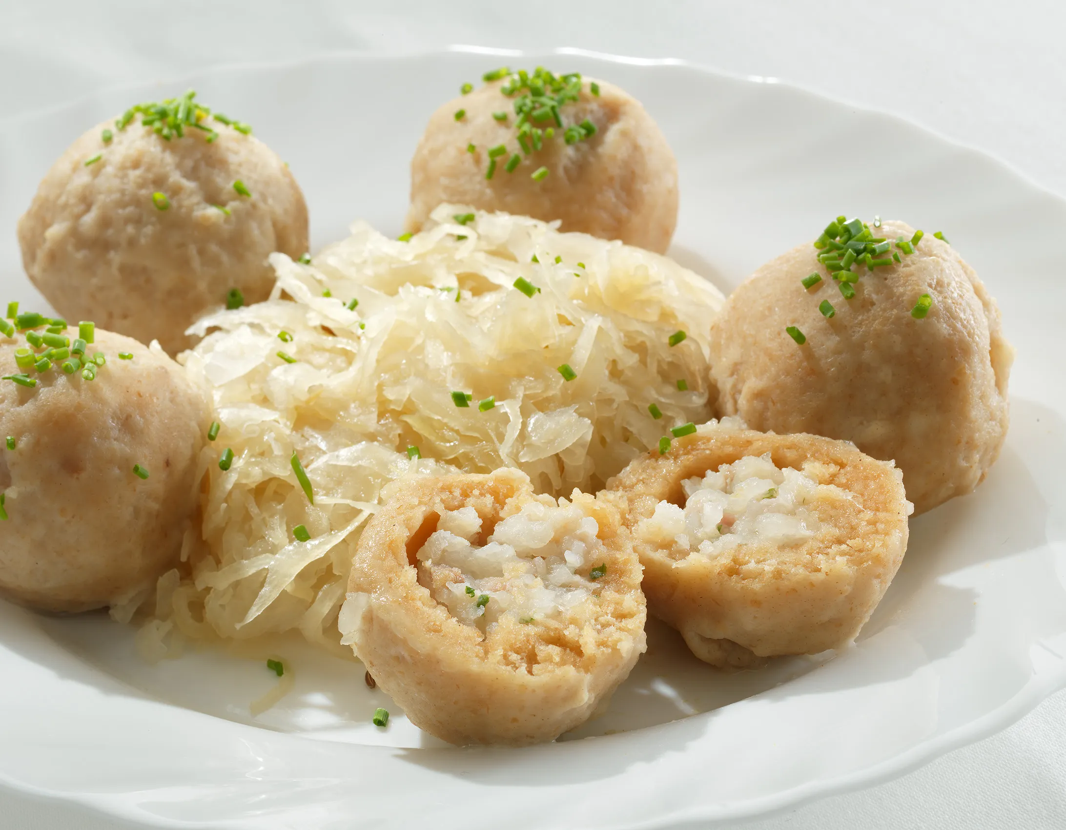 Auf einem weißen Teller liegen mehrere Knödel, in der Mitte Krautsalat. Im Vordergrund ist ein Knödel aufgeschnitten und hat eine helle Füllung.