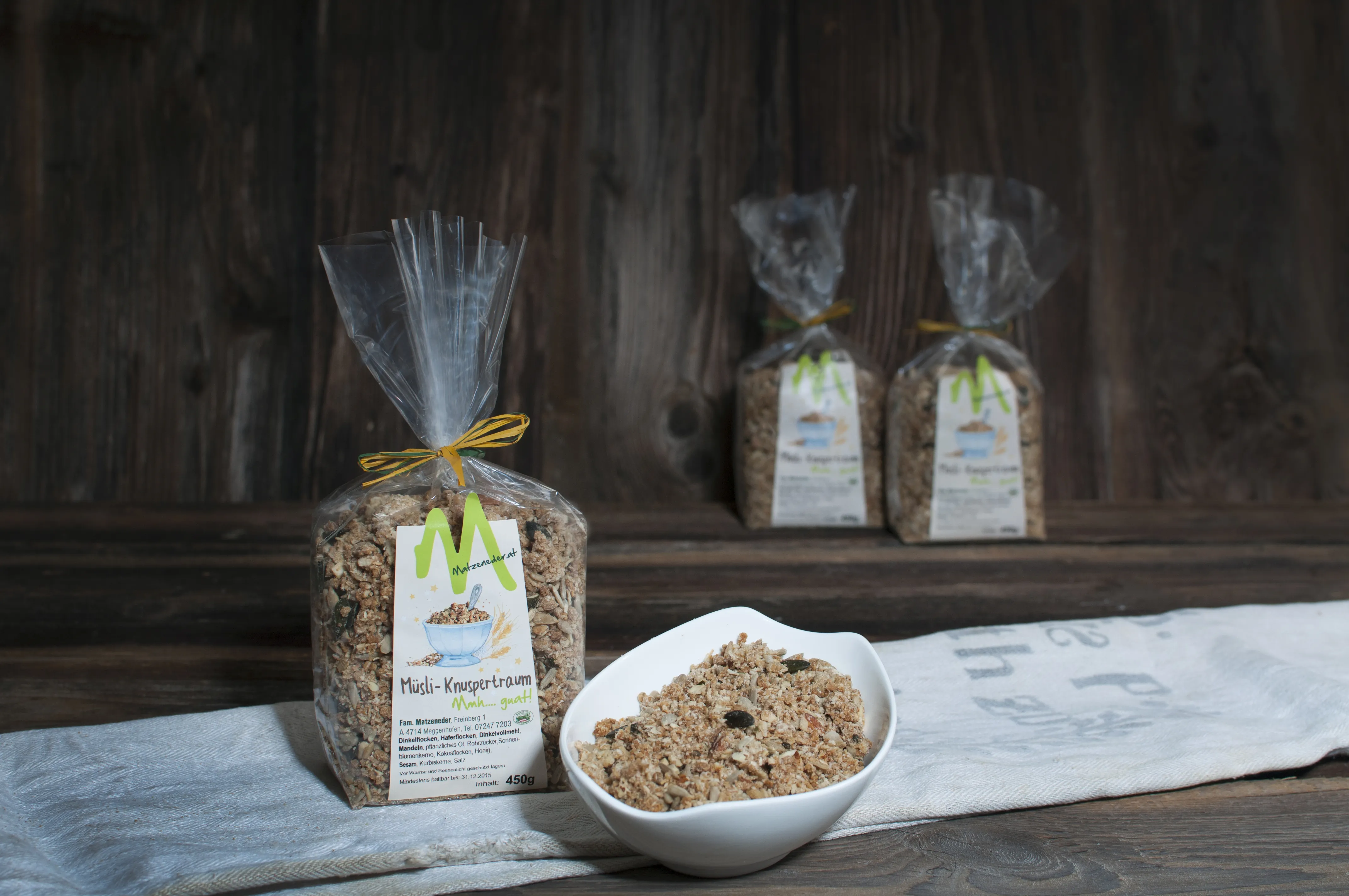 Auf dunklem Hintergrund liegt ein weißes Tuch, darauf steht eine Schale mit Müsli und ein Sackerl mit Müsli; dahinter zwei weitere Sackerl mit Müsli