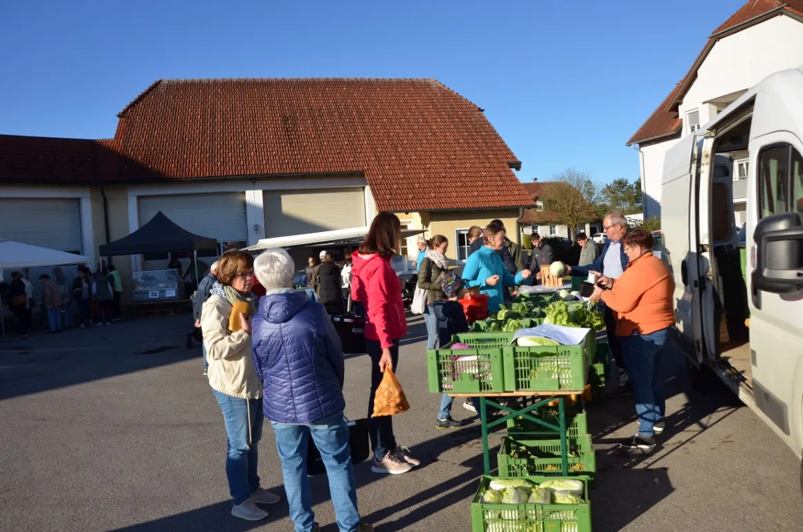 Man sieht verschiedene Personen an Marktständen, dahinter Gebäude und blauer Himmel