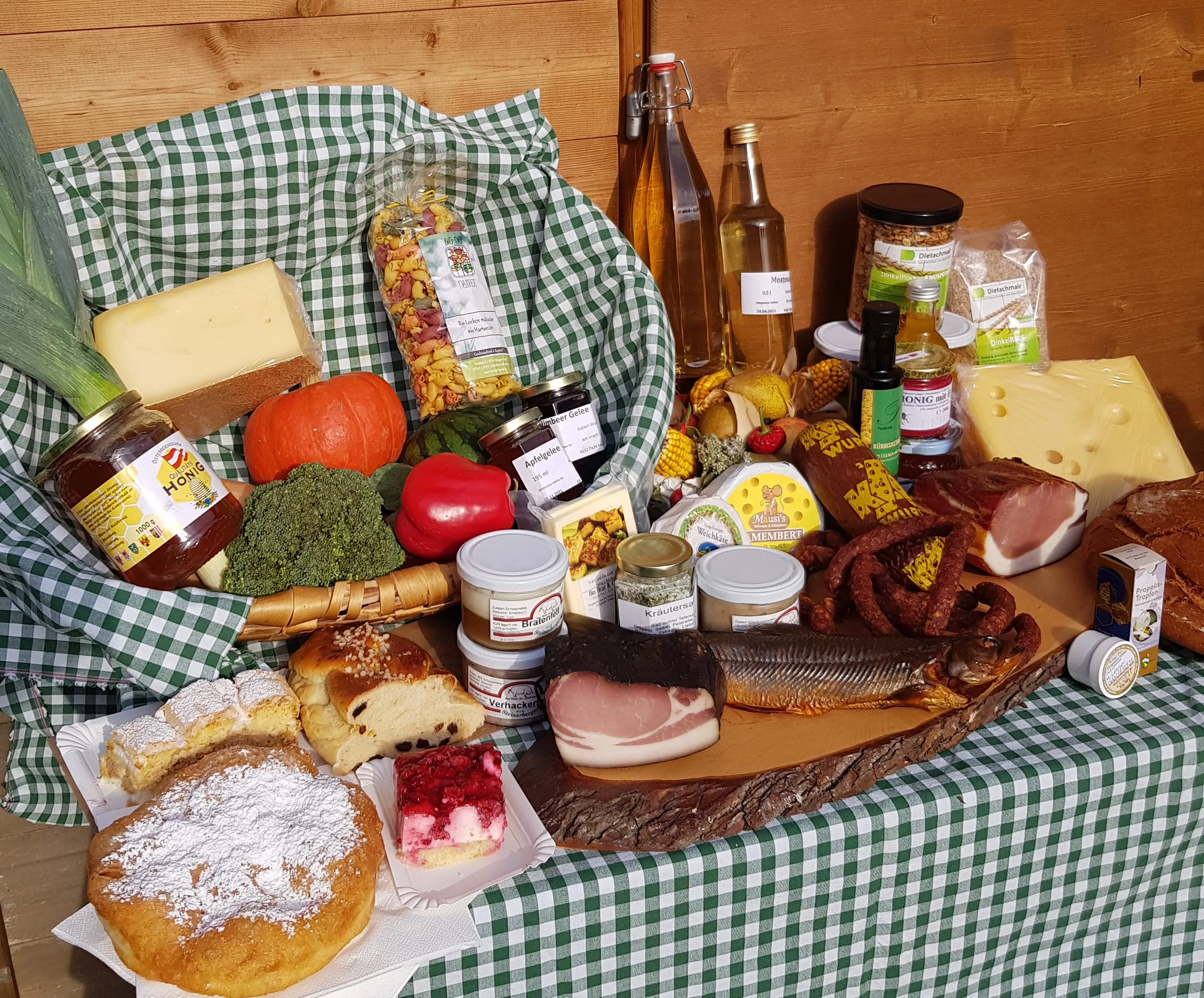 Man sieht einen Tisch, grün/weiß kariertes Tischtuch auf dem ein Korb, ebenfalls mit grün/weiß karierten Tuch steht. Auf dem Tisch und in dem Korb viele verschiedene Produkte wie Käse, Speck, Honig, Gemüse, Gläser, Flaschen und Mehlspeisen.