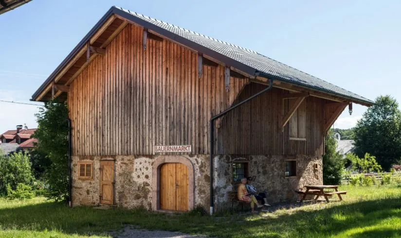 Auf dem Bild sieht man einen Holzstadel, mit zwei Türen, umgeben von grüner Wiese und dahinter blauer Himmel