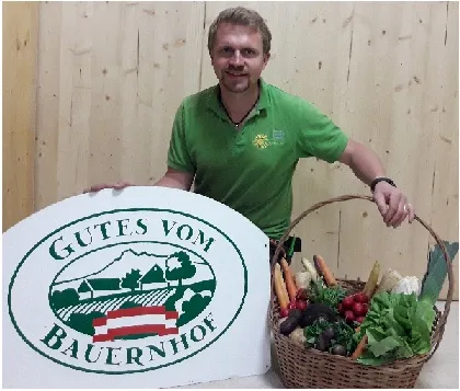 Herr Kirchgatterer steht vor einer Holzwand mit der Tafel Gutes vom Bauernhof in der einen Hand, in der anderen Hand hält er einen Korb mit verschiedenem Gemüse