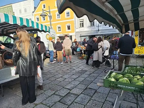 Man sieht mehrere Personen, welche auf einem Markt mit Ständen stehen, dahinter verschiedene Häuserfronten