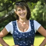 Romana Schneider-Lenz trägt ein weißes T-Shirt unter einem blauen, gemusterten Dirndl, steht mit Händen in der Hüfte im Freien.