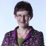 Maria Stütz mit kurzen, dunklen Haaren trägt eine lila gemusterte Jacke über einem grünen Oberteil und eine Halskette mit Kreuzanhänger.