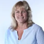 Elfriede Fuchs mit schulterlangem, gewelltem, blondem Haar trägt eine hellblaue Bluse mit offenem Kragen.