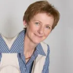 Maria Thek trägt blau-weiß gestreiftes Hemd und weiße Weste vor grauem Hintergrund.