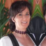 Monika Lindbichler-Sohneg mit schwarzem, kariertem Halsband und weißer Bluse vor grünen Pflanzen und orangefarbenem Hintergrund.